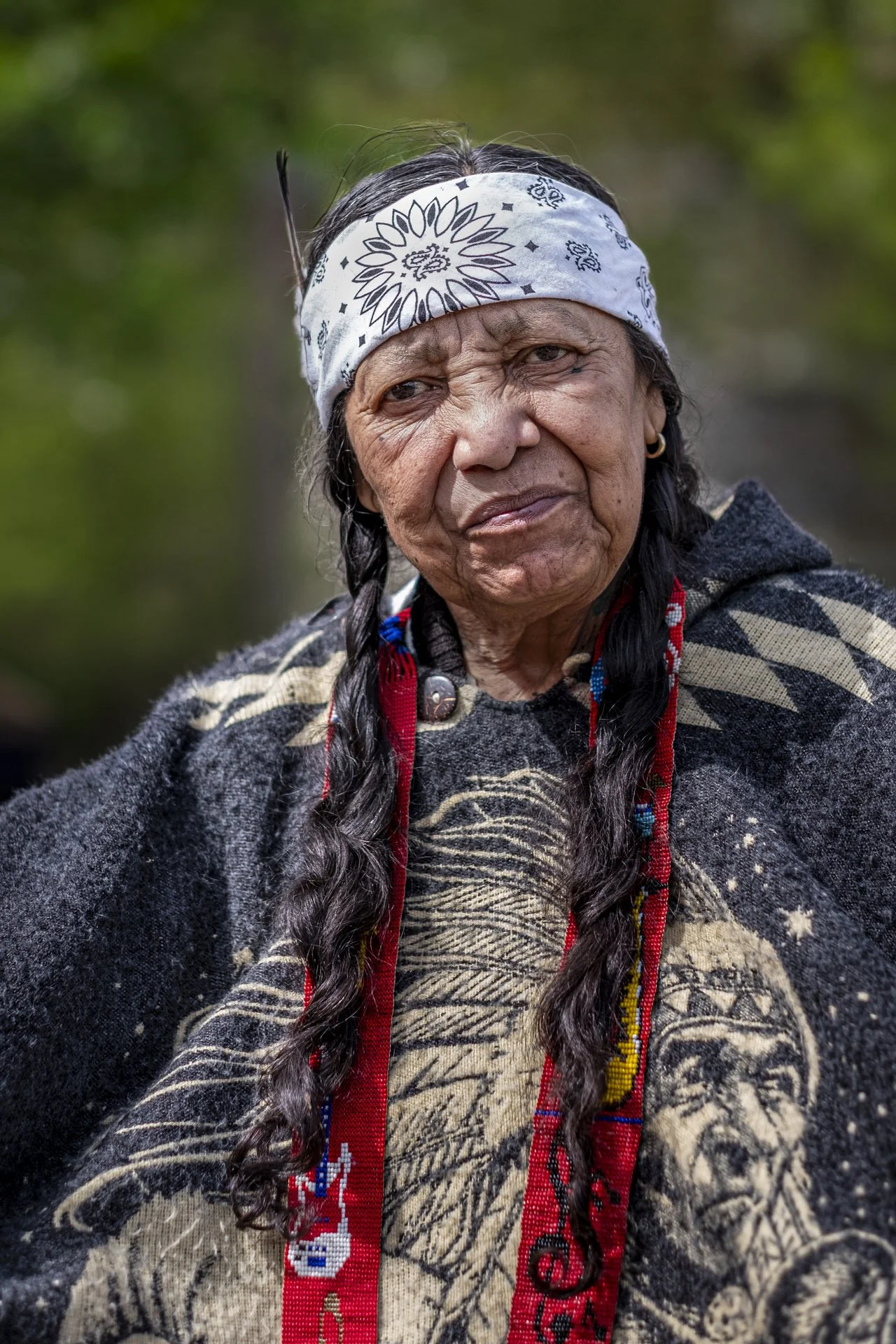 Close-up of an elderly Native American woman with long braided hair, wearing a white bandana with black patterns, a woven patterned garment, and traditional jewelry, outdoors with blurred green background.