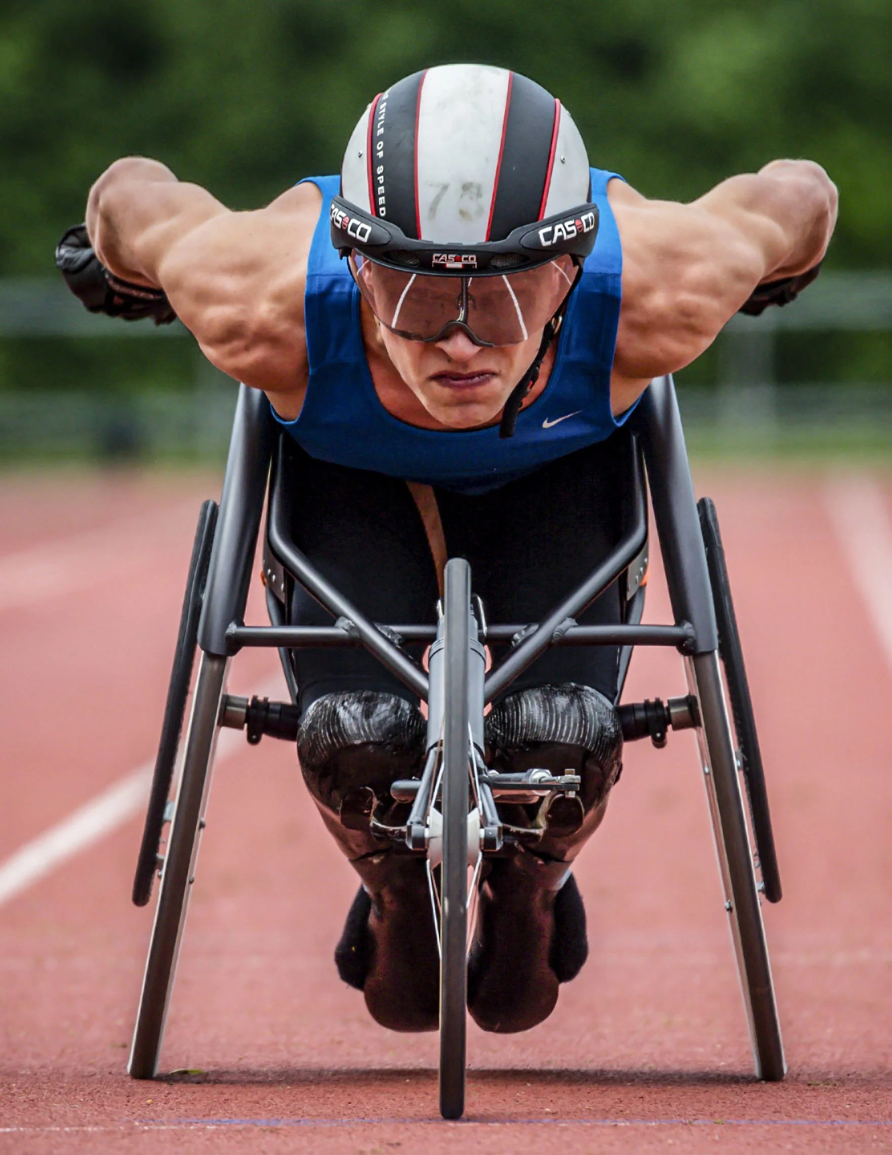 A male athlete in a wheelchair, preparing for a race with his head down, wearing a helmet and sports gear on a red running track.