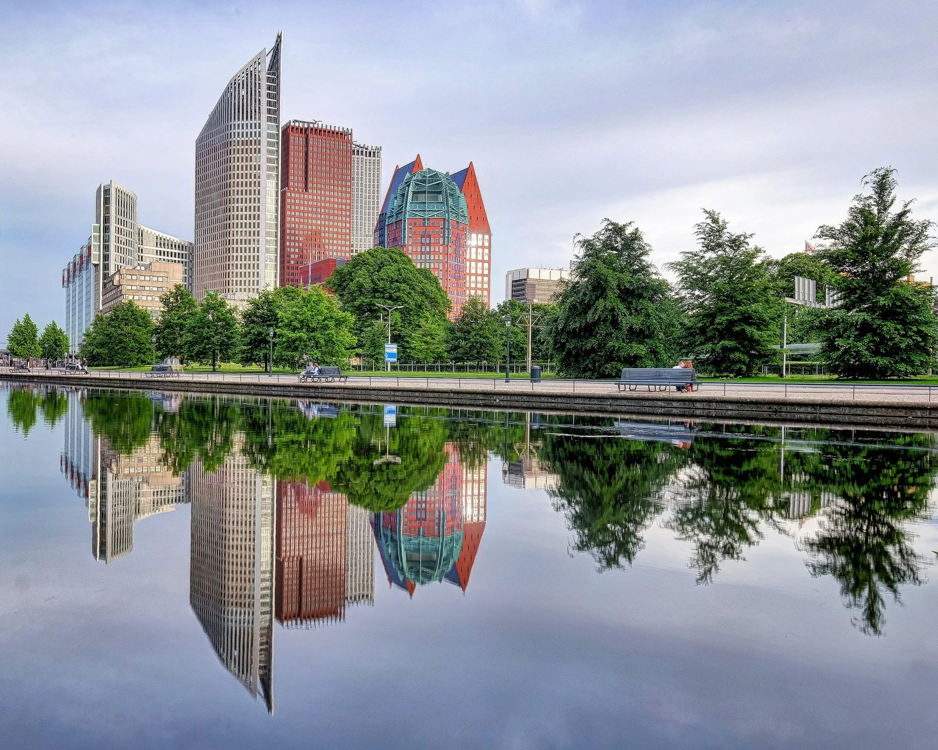 Reflection of a city skyline with tall buildings and trees along a riverbank, under a cloudy sky.