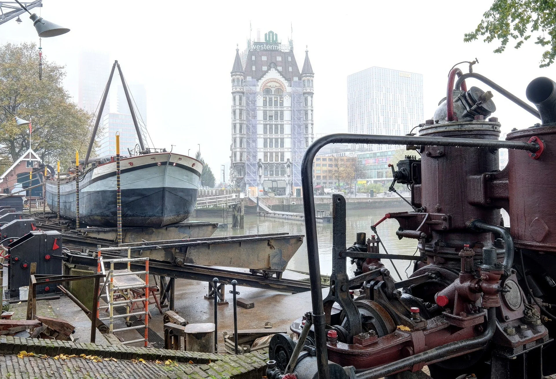 View of a historic boat docked at a waterfront with City Center buildings in the background, including a tall building with a clock tower and a structure with a sign reading 'Westermann'. An old red steam engine is visible in the foreground, and the 