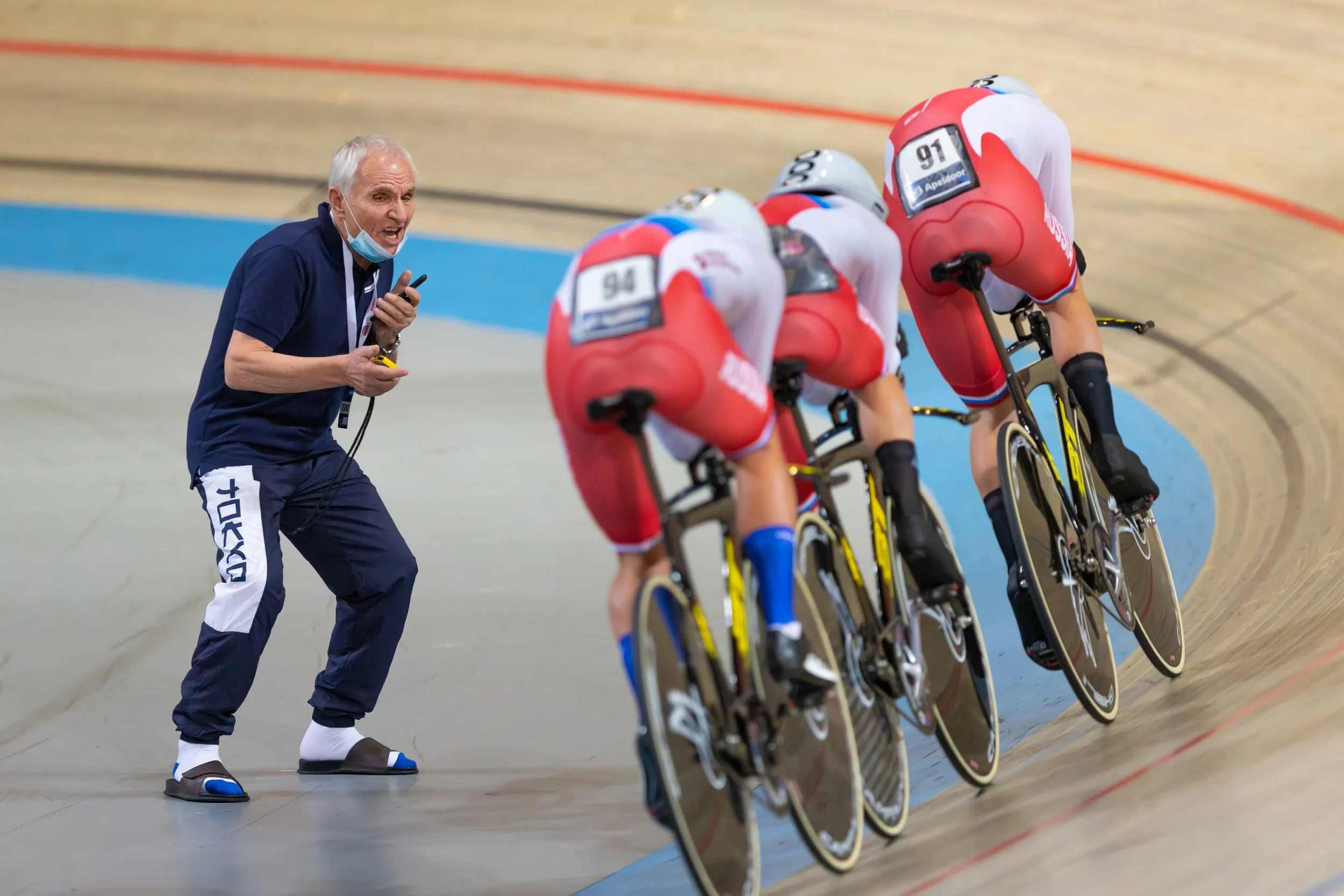 A cycling coach or team staff member in a navy blue shirt and white pants, with a face mask below his chin, is holding a stopwatch and speaking to a group of four speedskaters in a velodrome. The skaters are wearing matching red, white, and blue unif