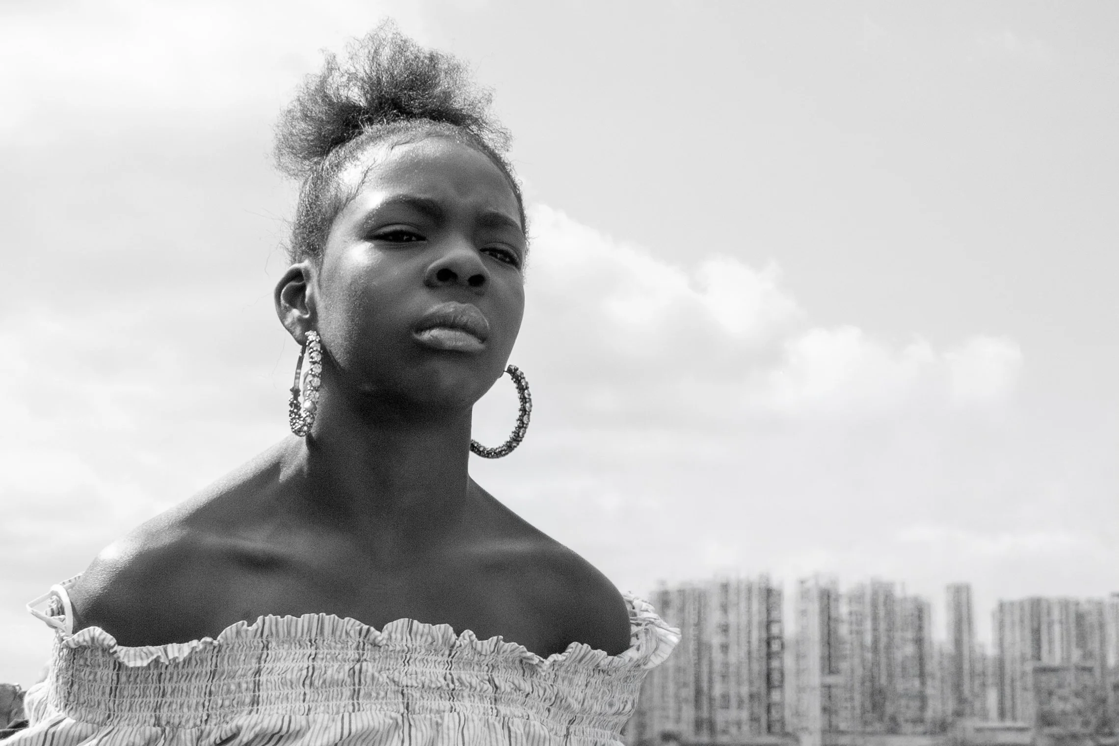 Black-and-white portrait of a woman with her hair styled in a high, curly puff, wearing large hoop earrings and a ruffled off-the-shoulder top, standing outdoors with a cloudy sky and cityscape in the background.
