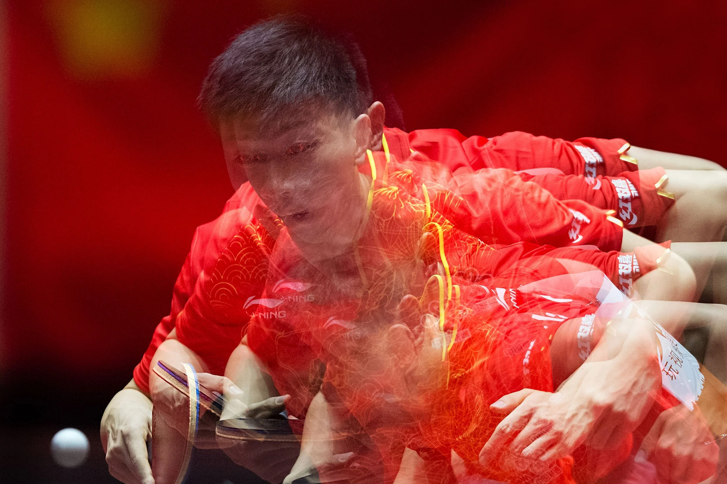 A male athlete in a red sports uniform sitting at a table, signing or writing in a book or paper, with a focused expression, with a dark background.