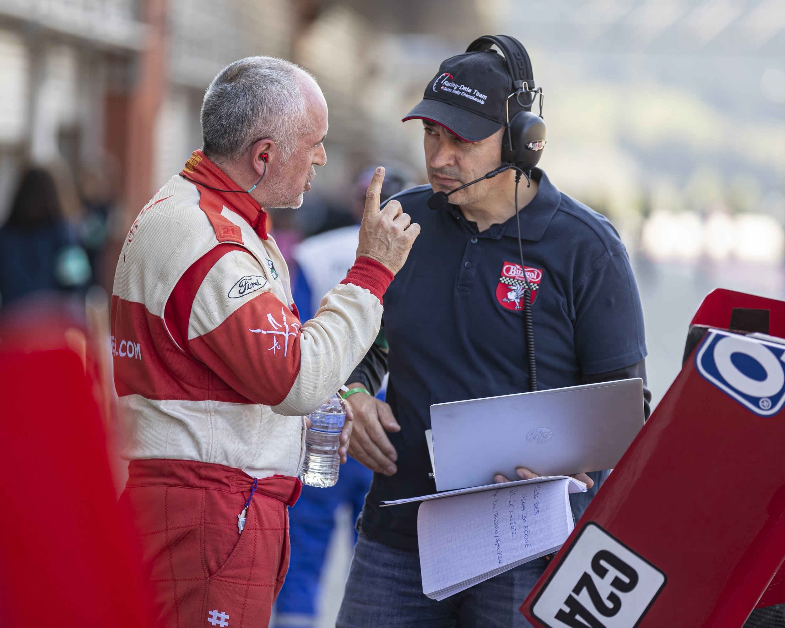 Two men are having a conversation at a racing event. One man is dressed in a racing suit with sponsor logos, and the other man is in a dark polo shirt with a headset, holding documents. There are signs and blurred spectators in the background.
