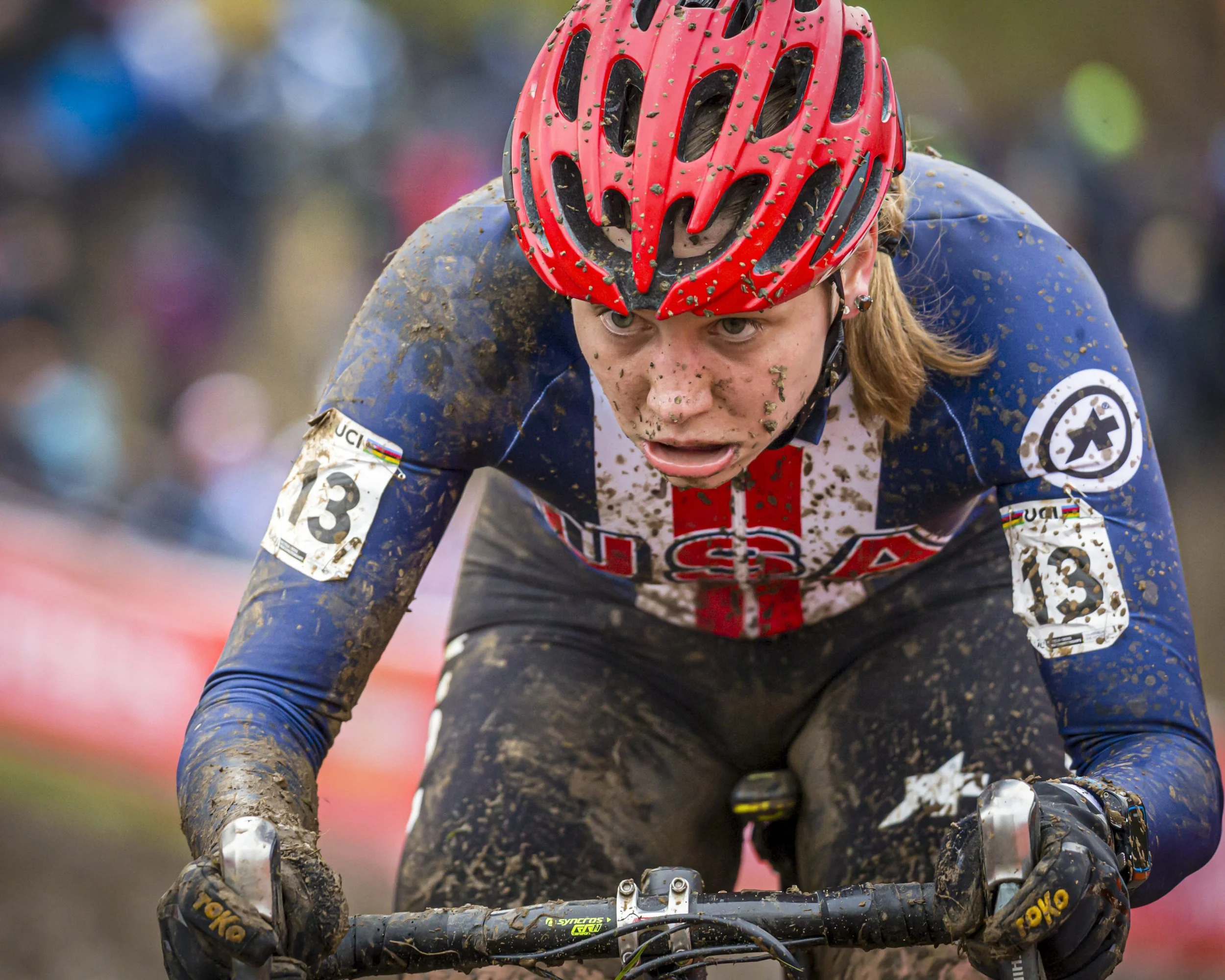 A cyclist wearing a red helmet and a racing jersey is riding on a muddy trail, covered in dirt and mud, with a focused expression on their face.