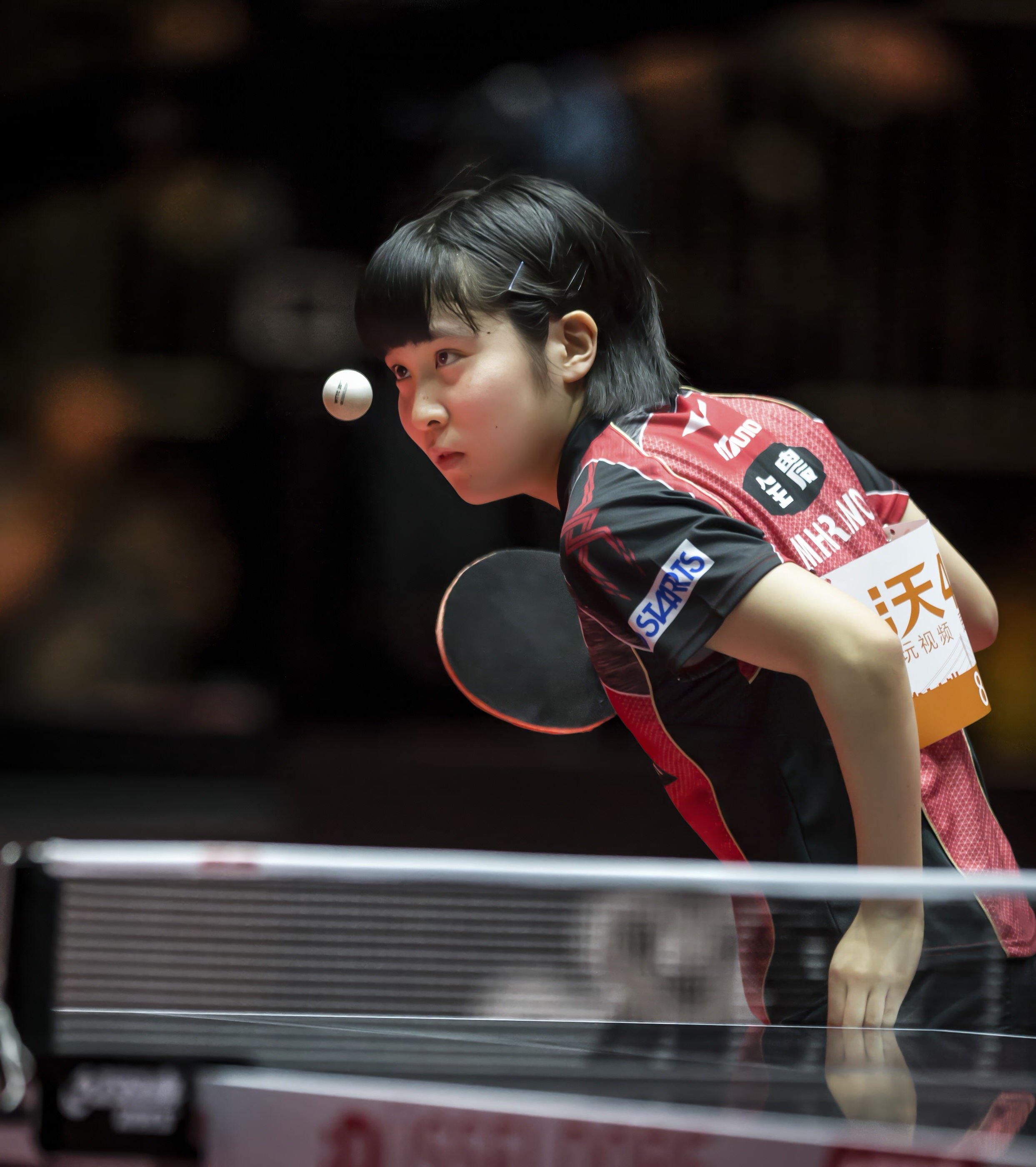 A female table tennis player with short dark hair in a red and black uniform, leaning forward with a focused expression, preparing to hit the ball during a match.