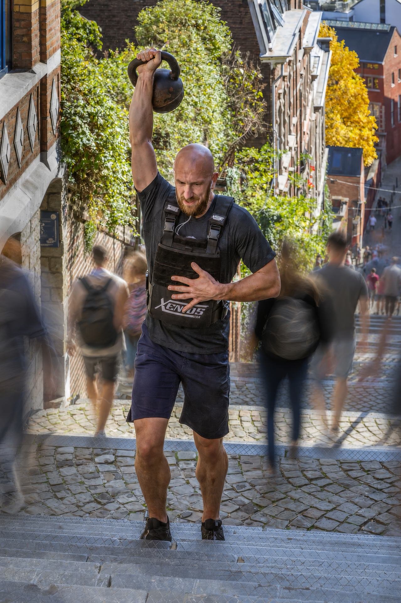 A man in black athletic clothing, holding a kettlebell overhead with one arm and a hand on his chest, is running up cobblestone stairs outdoors, surrounded by blurred pedestrians, with brick buildings and yellow-leaved trees in the background.