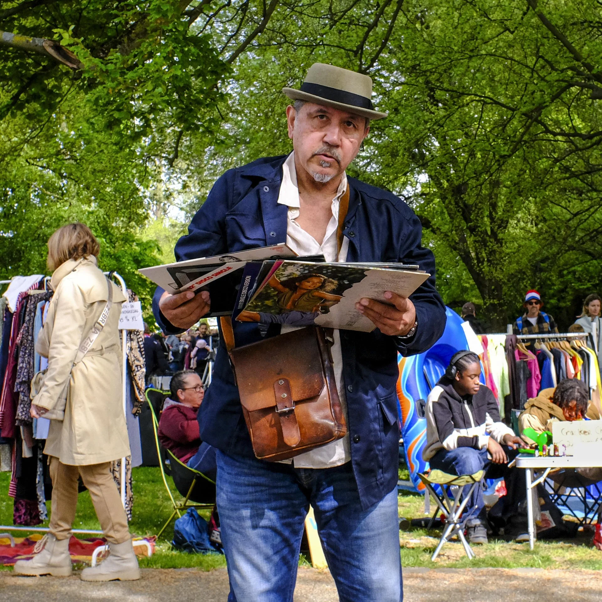A man with a beige fedora hat, light gray dress shirt, and dark jacket, holding a magazine, standing outdoors among people and market stalls with clothes and children in the background, under green leafy trees.