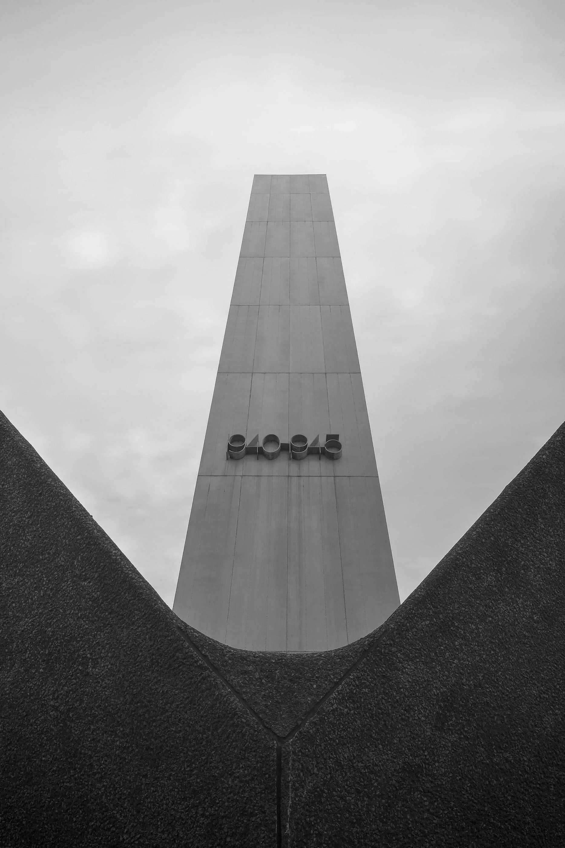 A black and white photo of a tall monument or obelisk viewed from below, with the sky and clouds in the background and the image framed by dark textured borders.