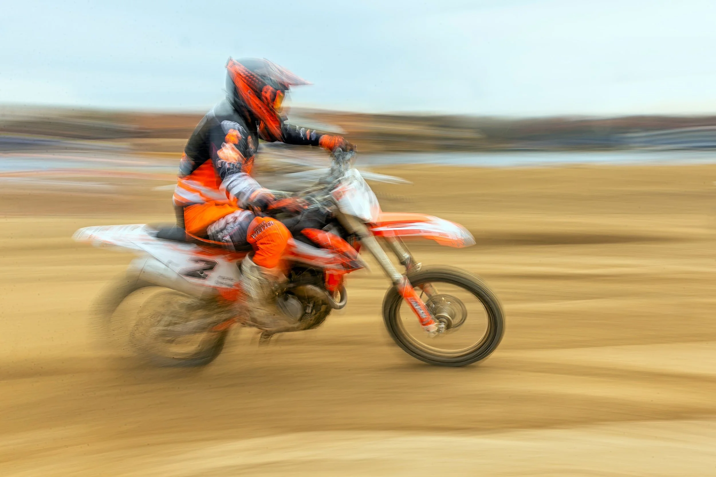 A person wearing a helmet and protective gear riding an orange dirt bike on a dirt track with motion blur.
