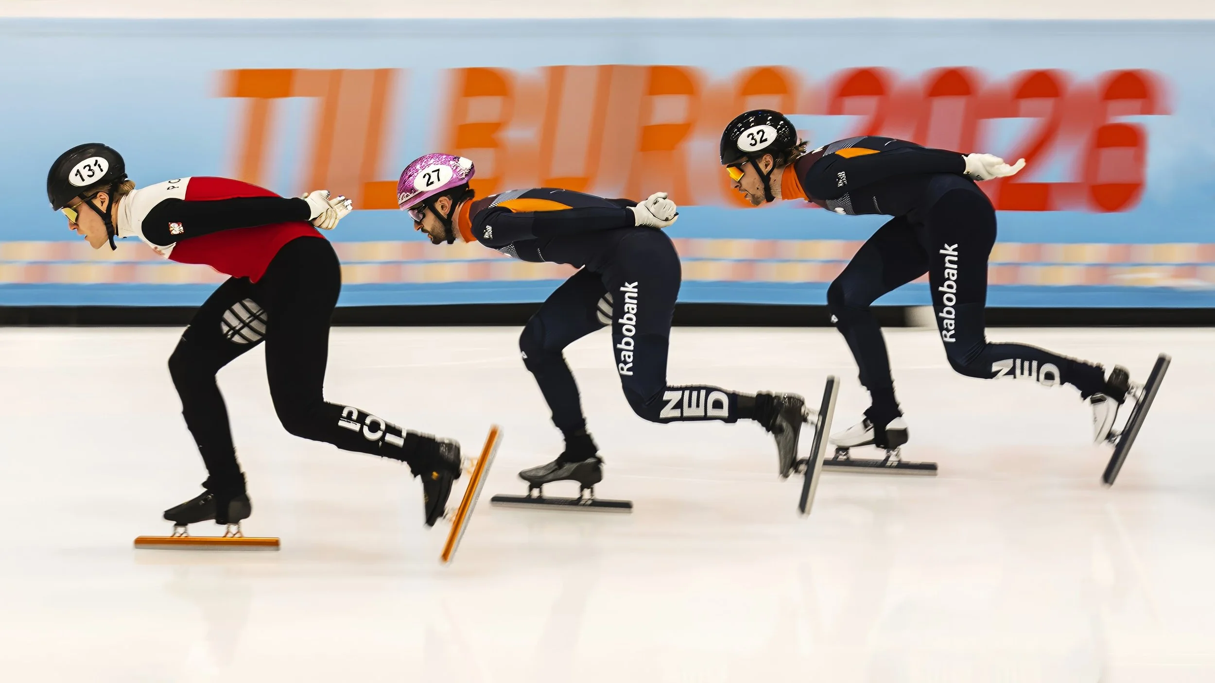 Three speed skaters in a race, wearing helmets and athletic gear, leaning forward on an indoor ice rink.