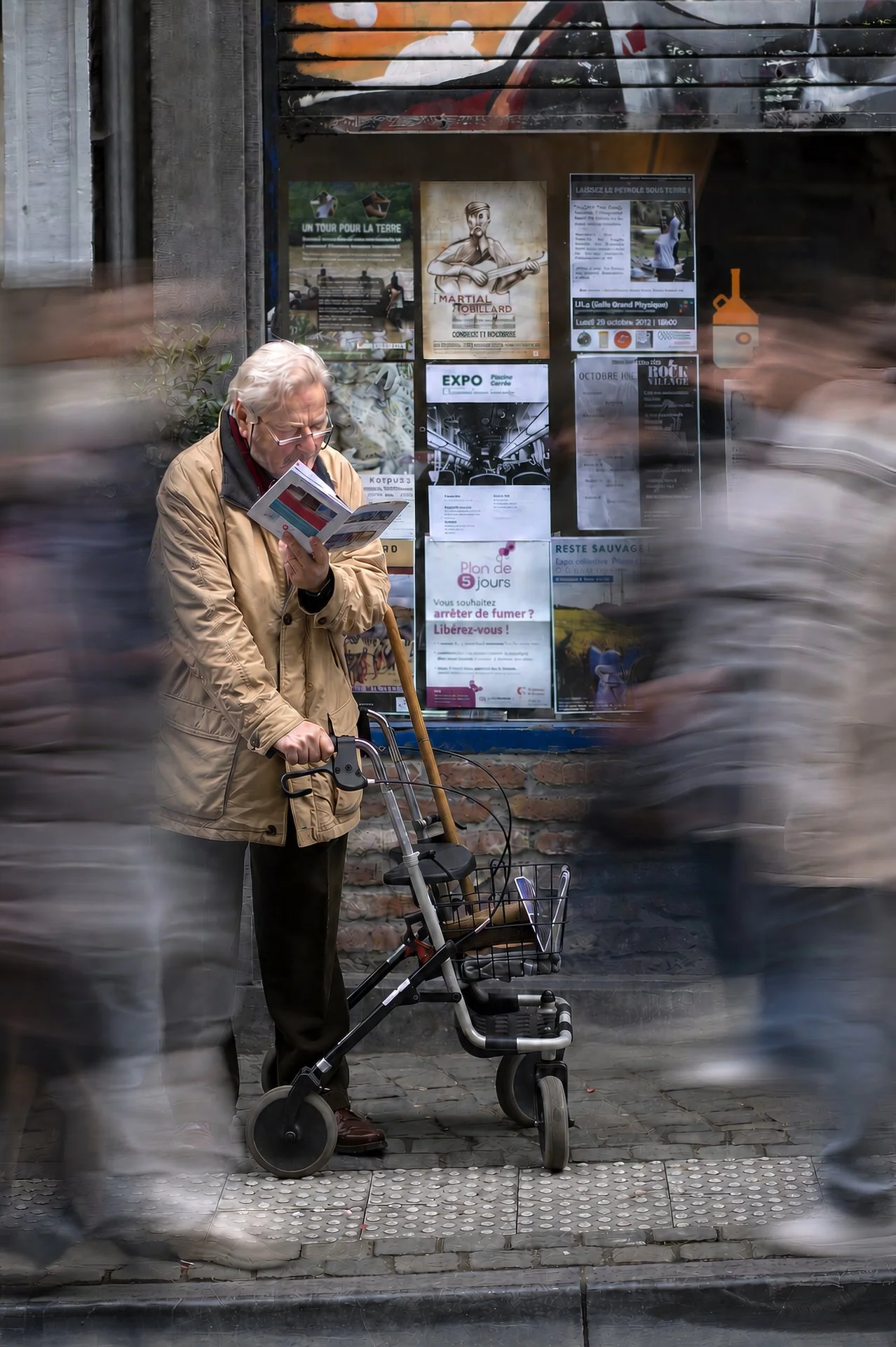 An elderly man with gray hair, glasses, and a beige jacket stands on a city street, reading a pamphlet or flyer. He uses a walker with a basket and has a cane tucked under his arm. The background shows a wall with various posters and flyers, a small 