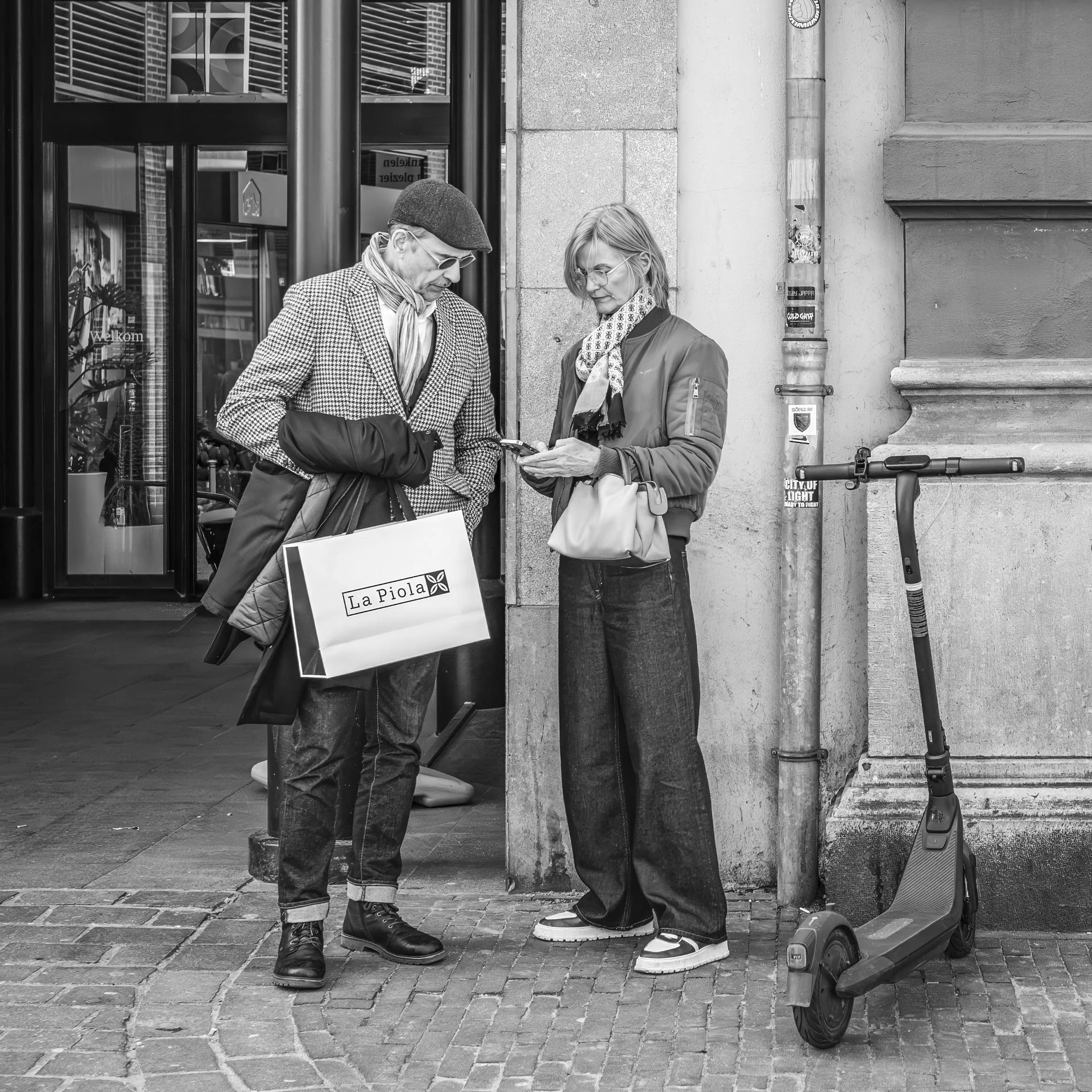Two people, a man and a woman, are standing on a cobblestone street outside a building, looking at a smartphone together. The man is wearing sunglasses, a beret, a checkered blazer, and gloves, and is holding a shopping bag. The woman has glasses, a 