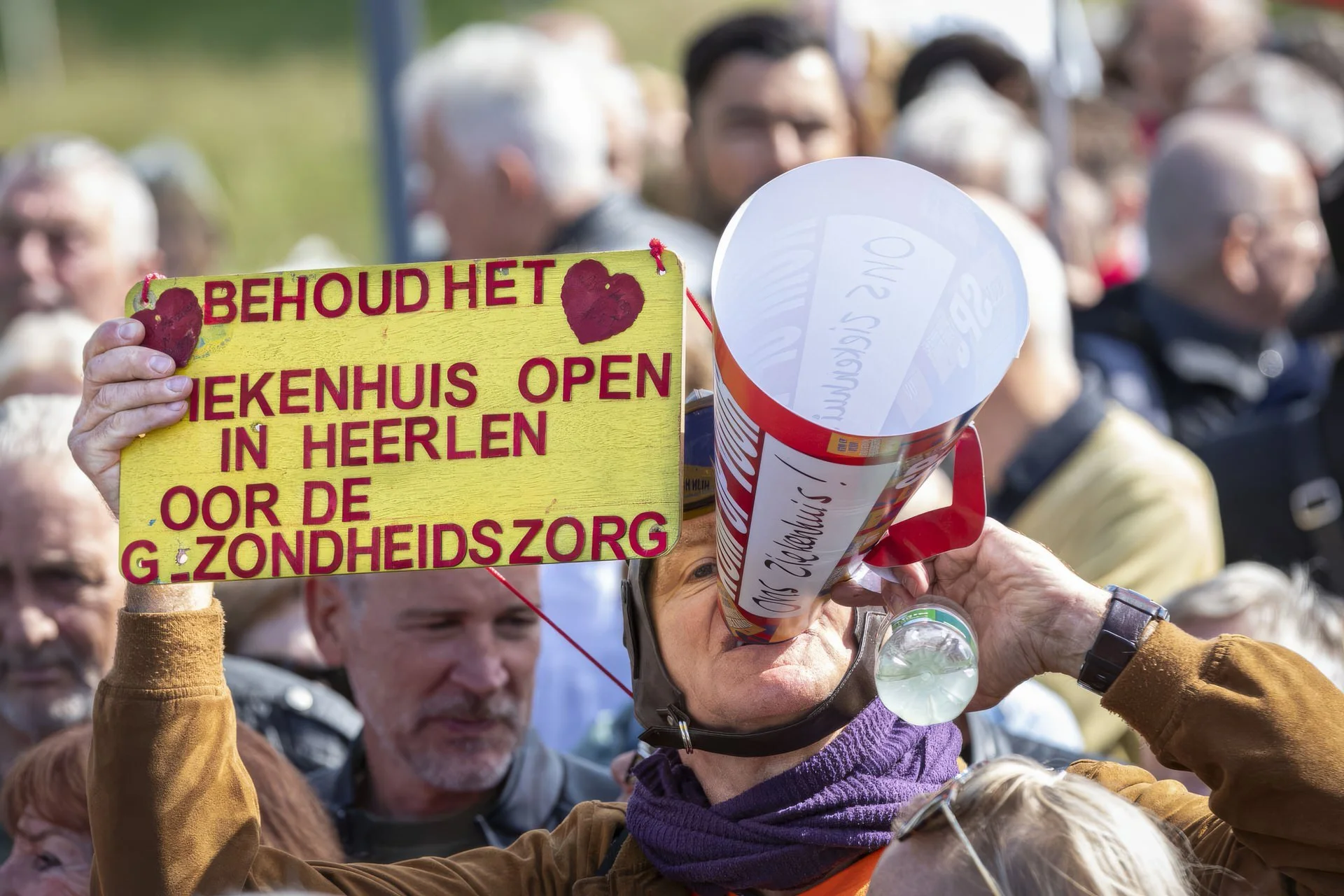 A group of people at a rally, holding signs and a megaphone, with a focus on a yellow sign with red Dutch text and hearts, and a woman's face partially visible through the megaphone.