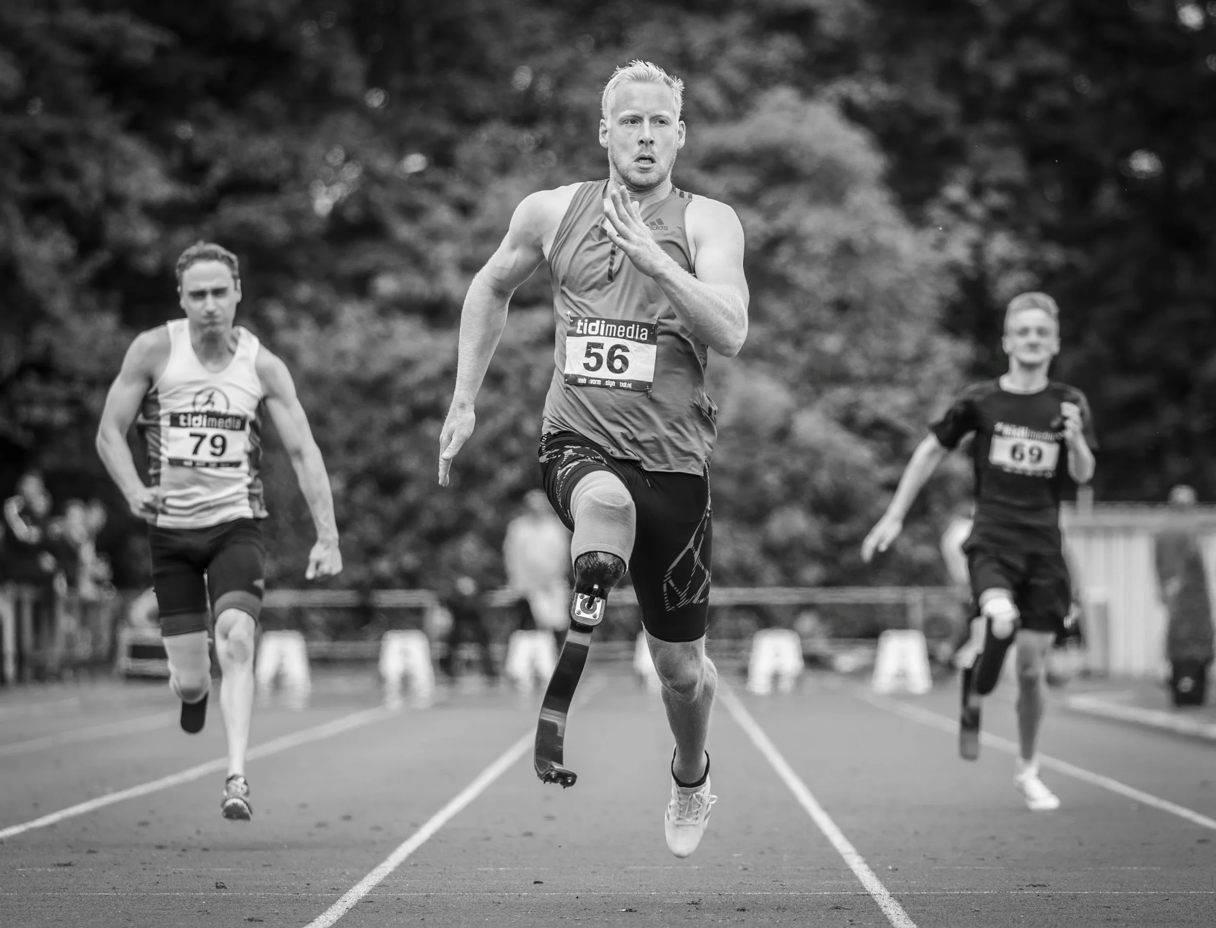Black and white photo of a male runner with a prosthetic leg competing in a race, with two female runners in the background, on a track with trees in the background.