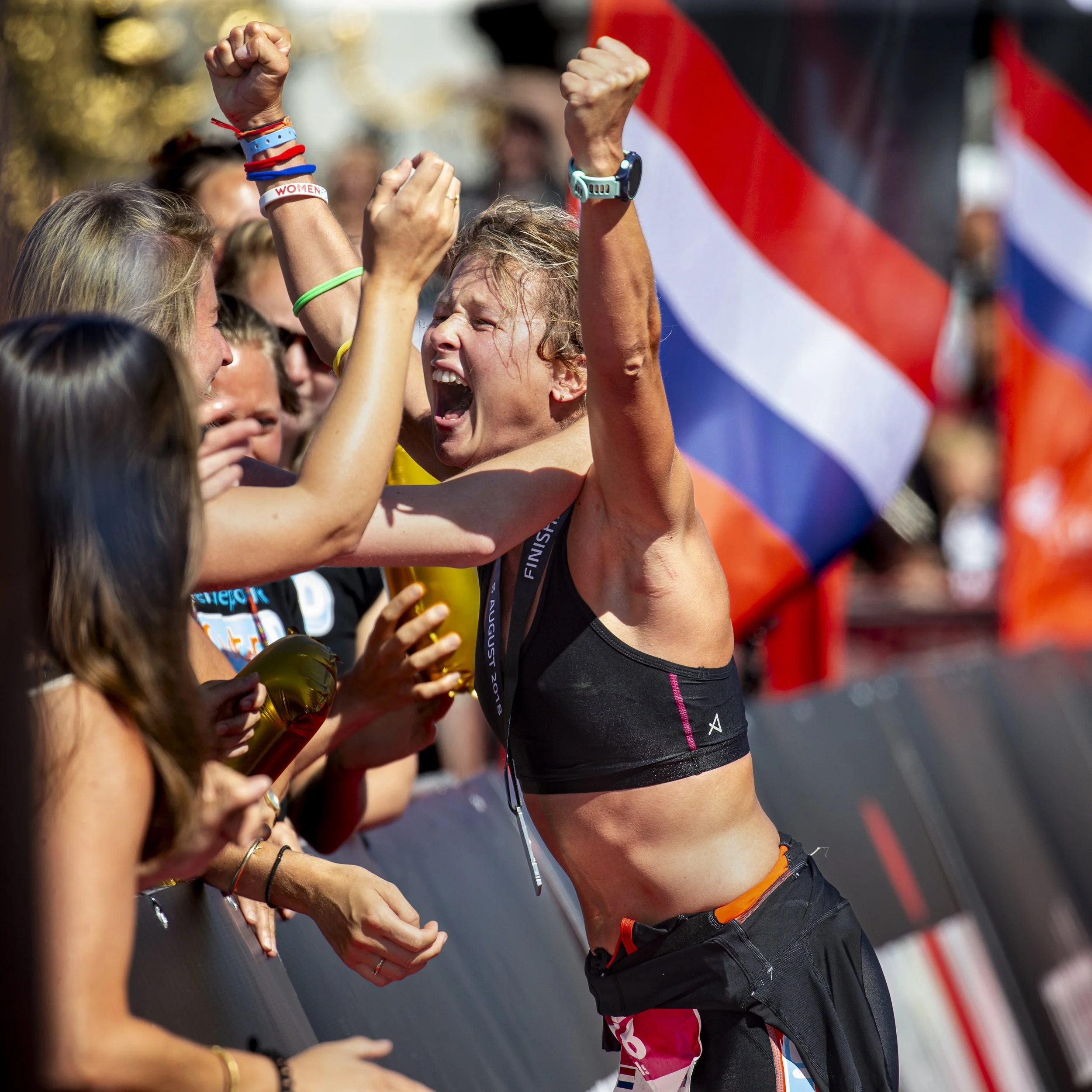 A woman celebrating her achievement at a race, raising her fists in the air, with friends or supporters around her. She is wearing a medal, weather-appropriate athletic gear, and various wristbands. The background shows a volunteer or official and pa