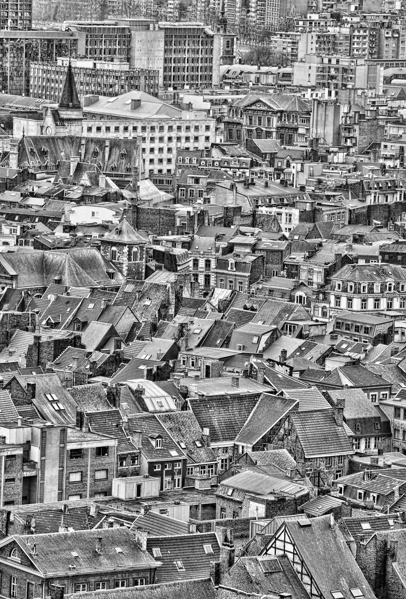 A black and white aerial view of a densely populated city with a mix of old and modern buildings, including rooftops, towers, and high-rise apartment complexes.