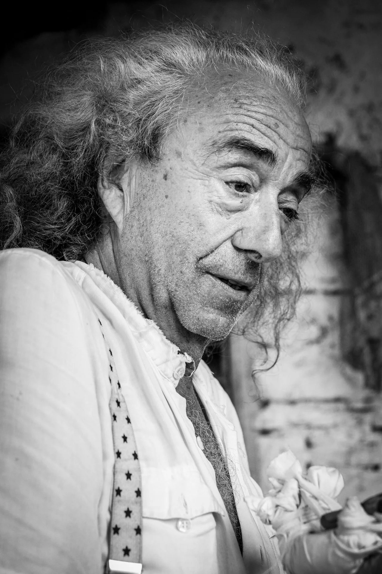 A black-and-white photo of an elderly man with long, curly hair, wearing a shirt with star-patterned suspenders, and holding a bunch of roses in his hand.