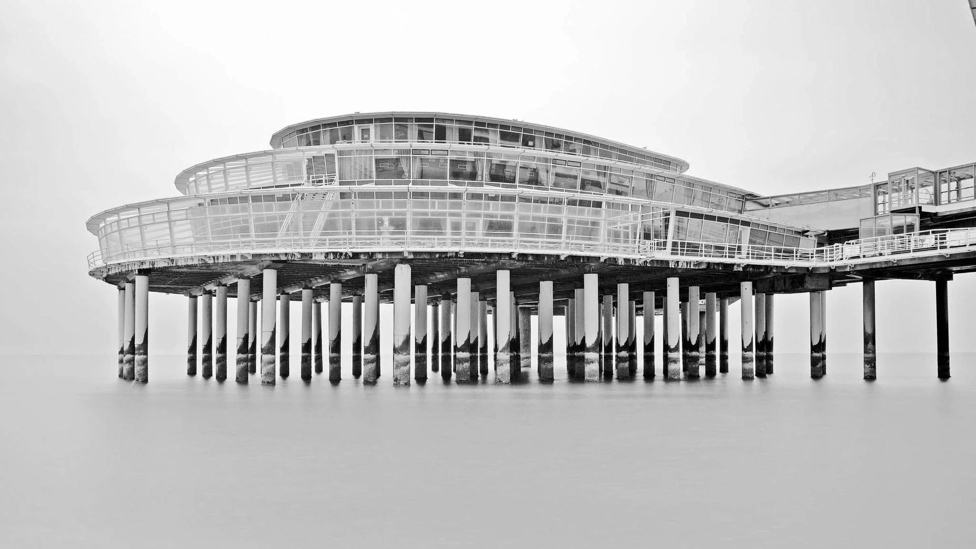 A black and white photo of a modern structure built on stilts over water, with curved glass walls and a white railing.