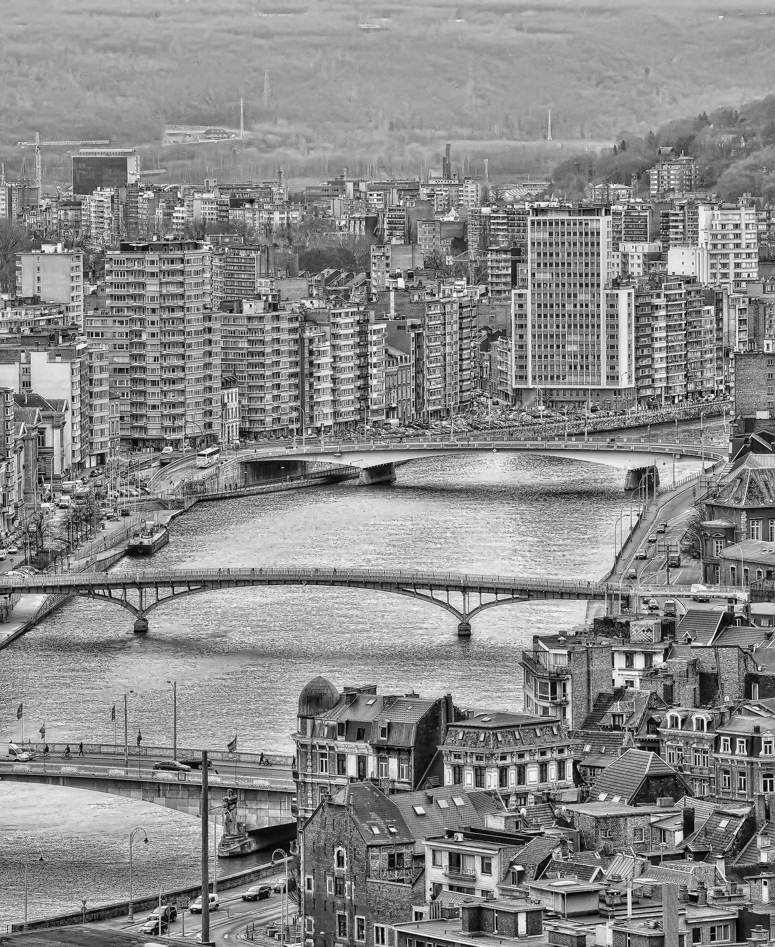 A black and white cityscape of a European city with tall buildings, bridges over a river, boats, and traffic.