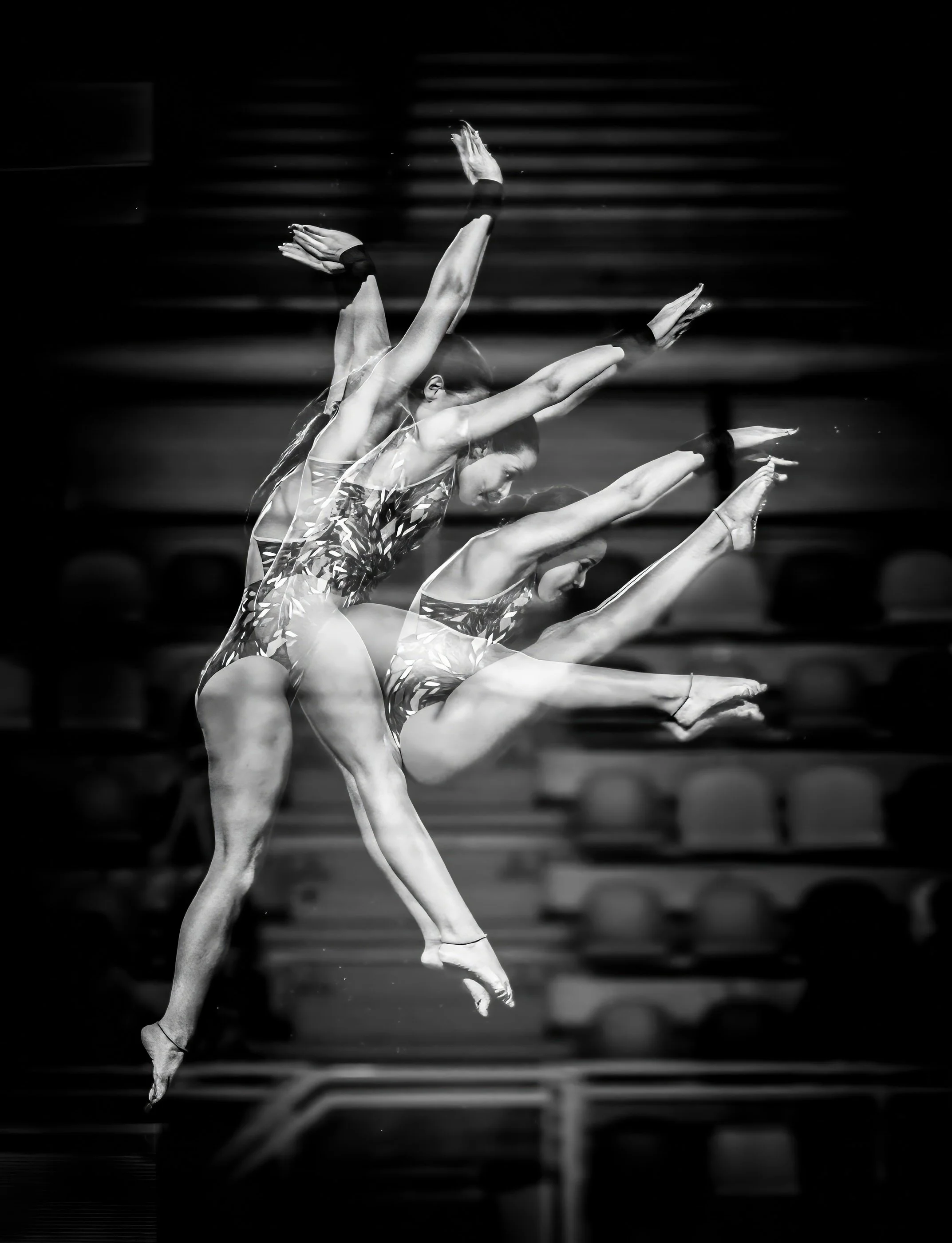 Black and white photo of three ballet dancers performing a synchronized leap on stage, with an out-of-focus set of stairs in the background.