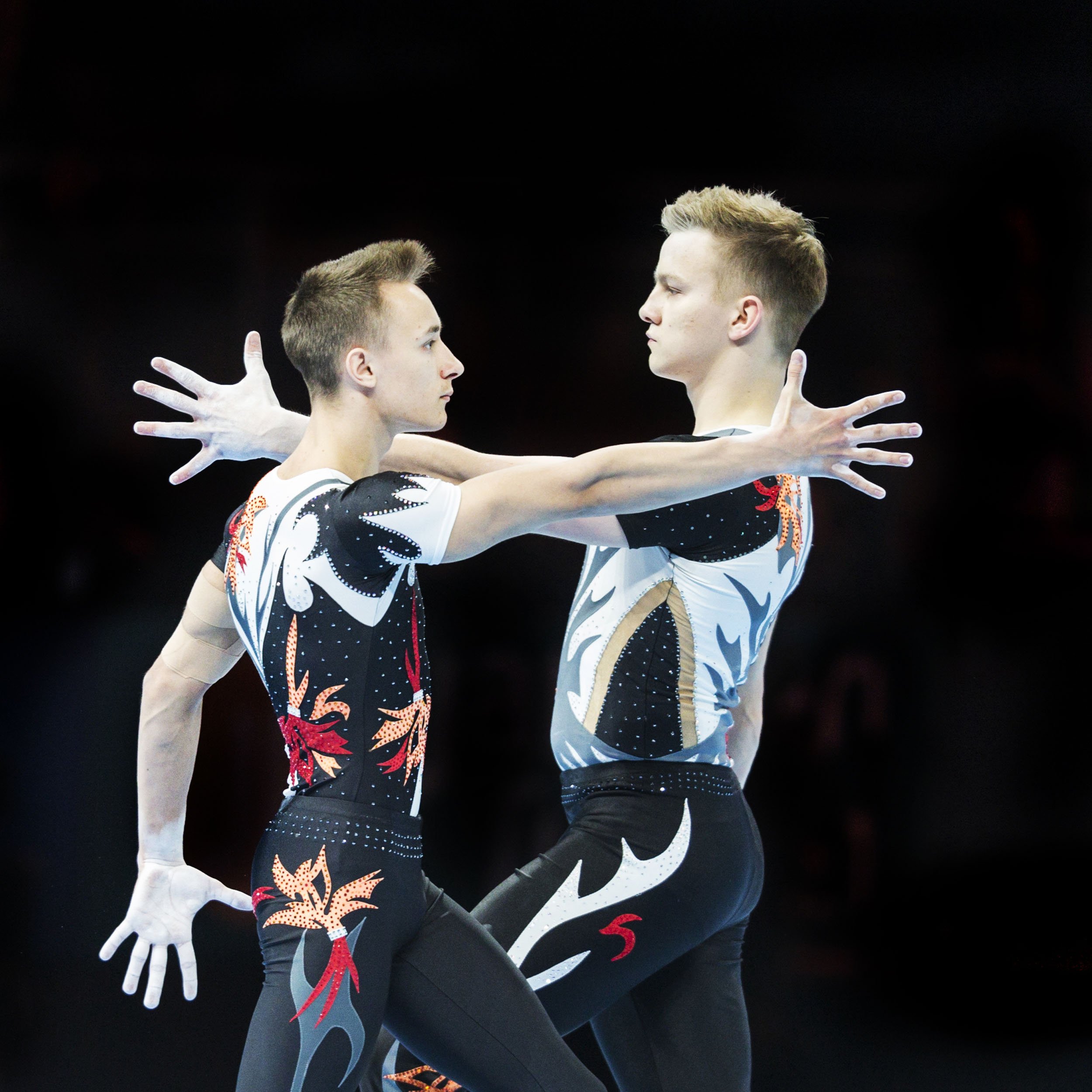 Two male gymnasts stretch their arms facing each other, with a black background.