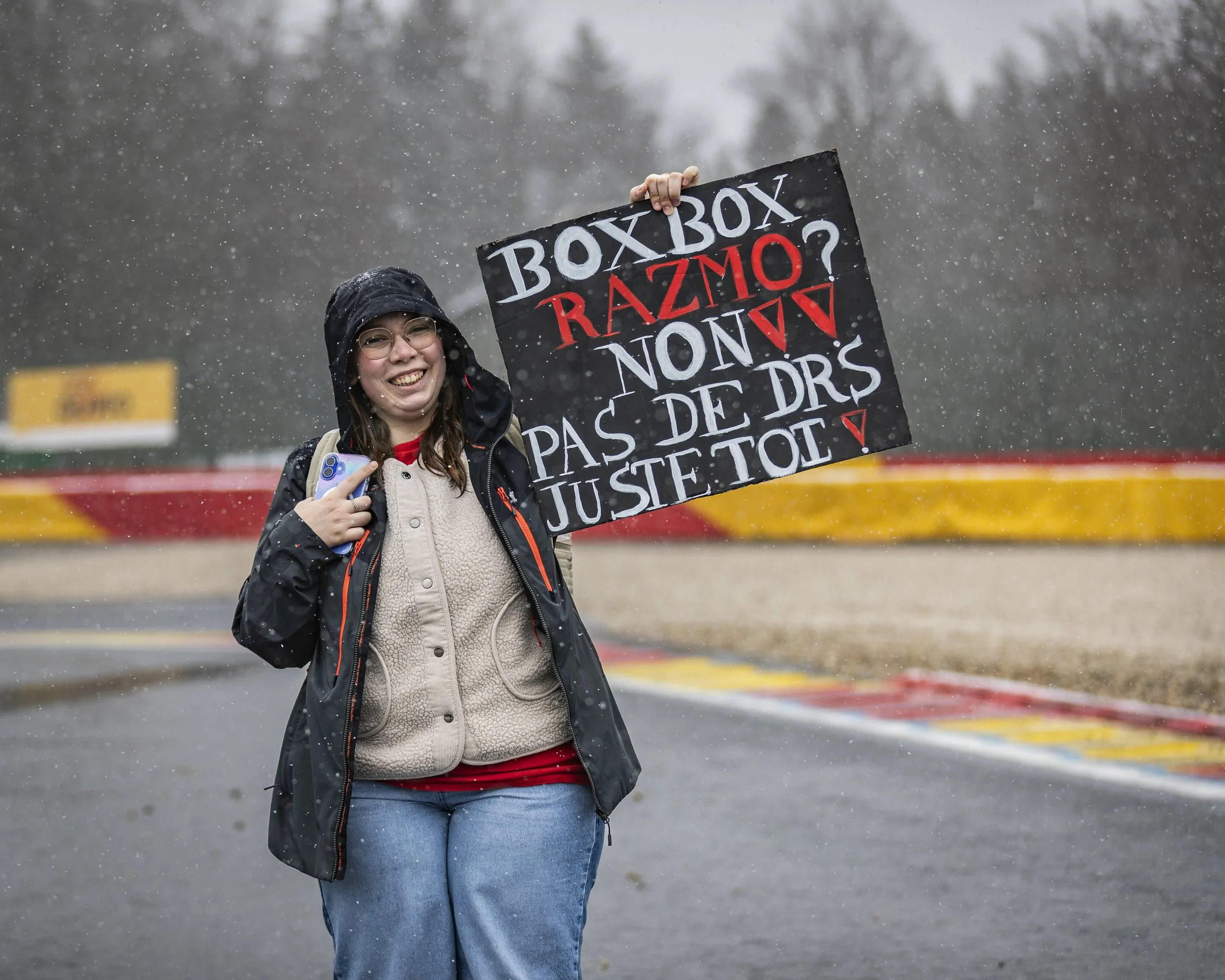 A smiling woman wearing glasses and a rain jacket holding a protest sign that reads, 'BOXBOX RAZMO? NON PAS DE DRS JUSTE TOL,' standing outside in rainy weather with trees and a road in the background.