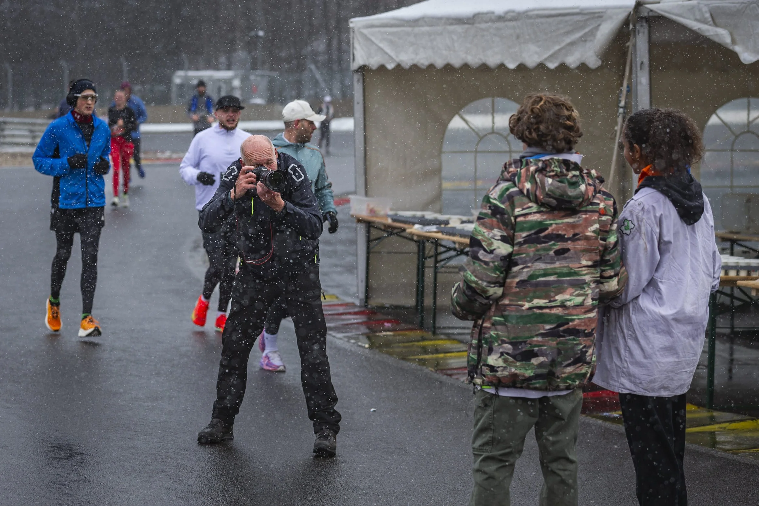 Runners participating in a race in rainy weather, with some spectators or officials standing by, in an outdoor setting with tents and wet pavement.
