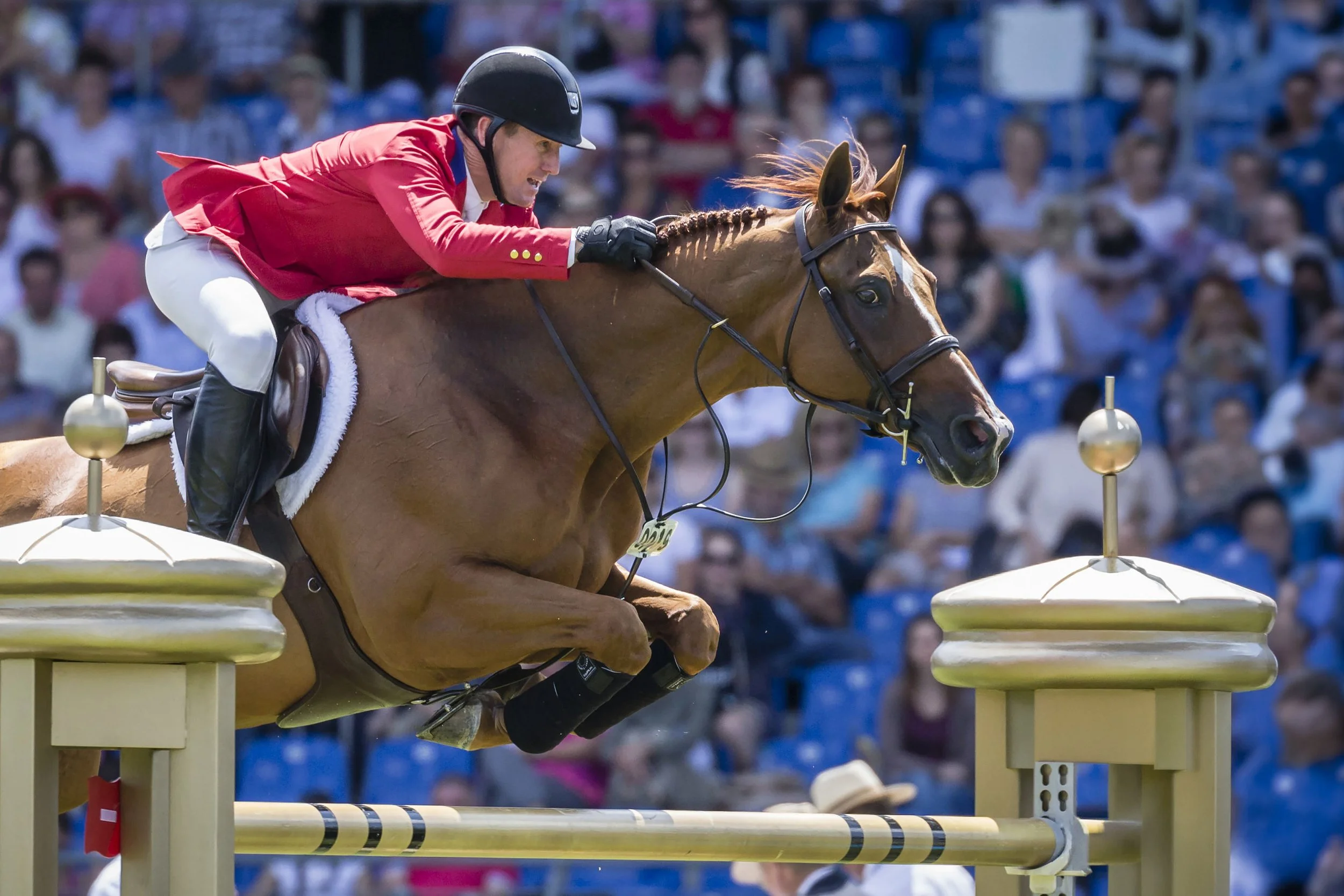 A male equestrian rider in a red jacket and black helmet riding a brown horse during a show jumping event, with the horse and rider jumping over a yellow hurdle.
