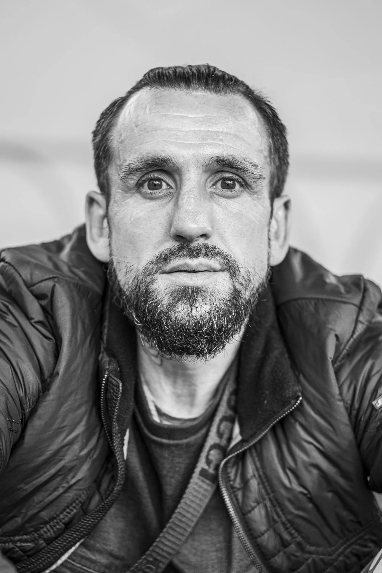 Black and white portrait of a man with a beard, mustache, and slicked-back hair, wearing a puffer jacket and looking directly at the camera.
