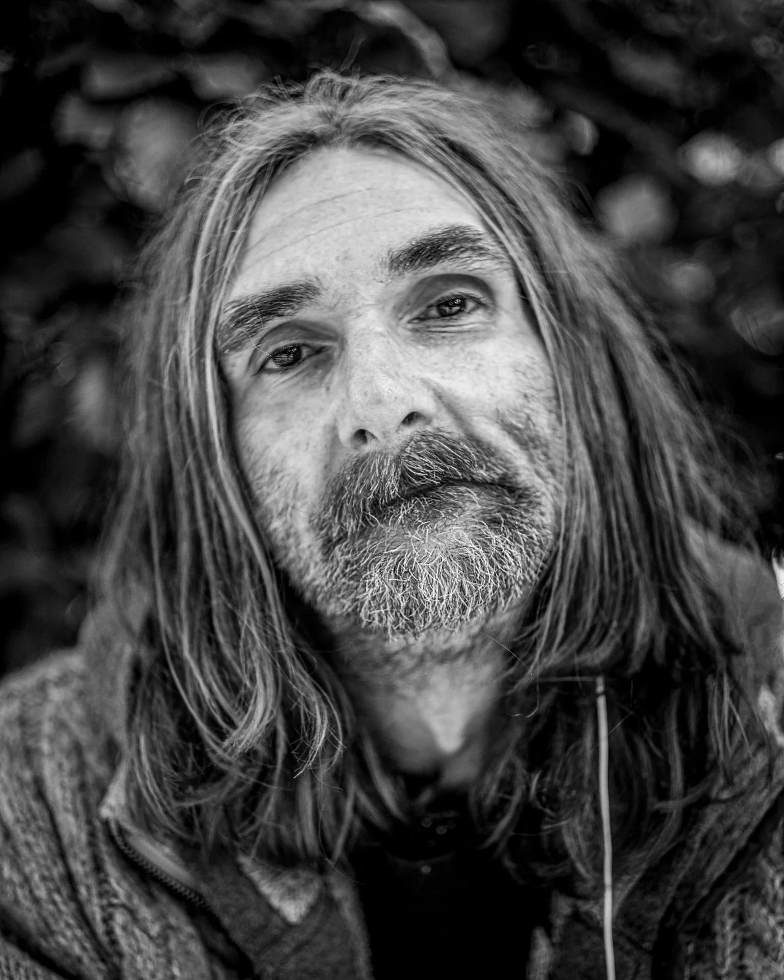 Close-up black and white portrait of a man with long hair, beard, and mustache, wearing a jacket with a fur collar.