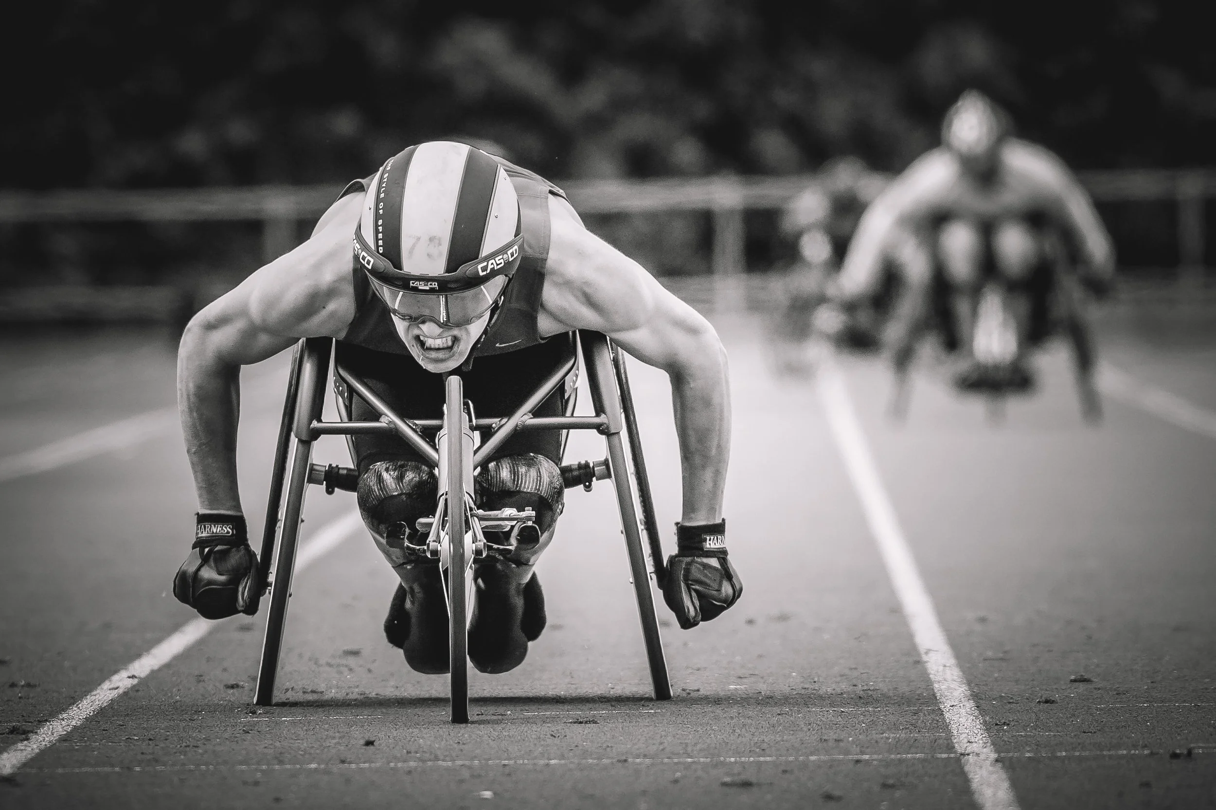 Black and white photograph of a male wheelchair racer in a crouched position on a track, wearing a helmet and sports glasses, with another wheelchair racer in the background.