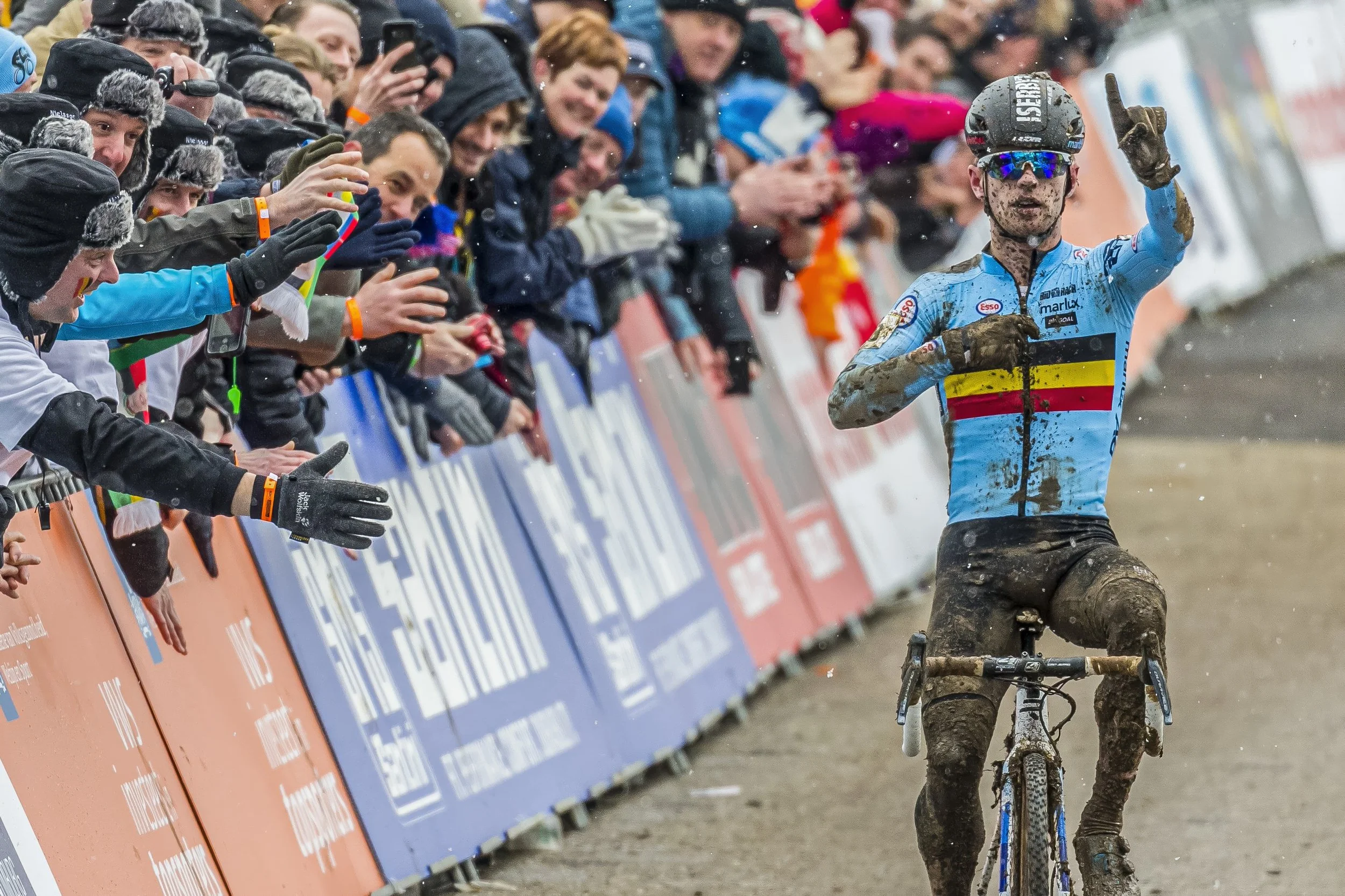 Mud-covered cyclist in a blue jersey celebrating victory on a muddy mountain bike race track, with cheering spectators behind a barricade.