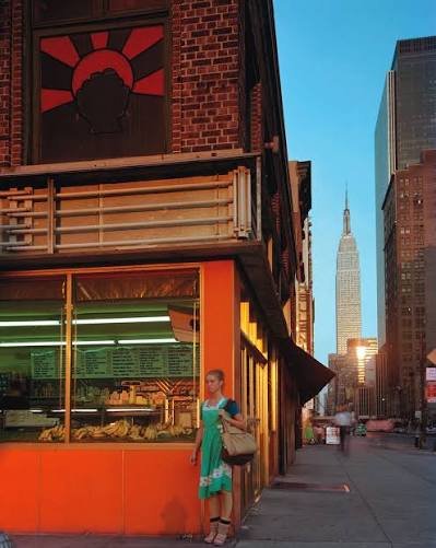 A woman standing on a city sidewalk with a bag, next to a store with large windows, in front of a brick building. The Empire State Building is visible in the background.