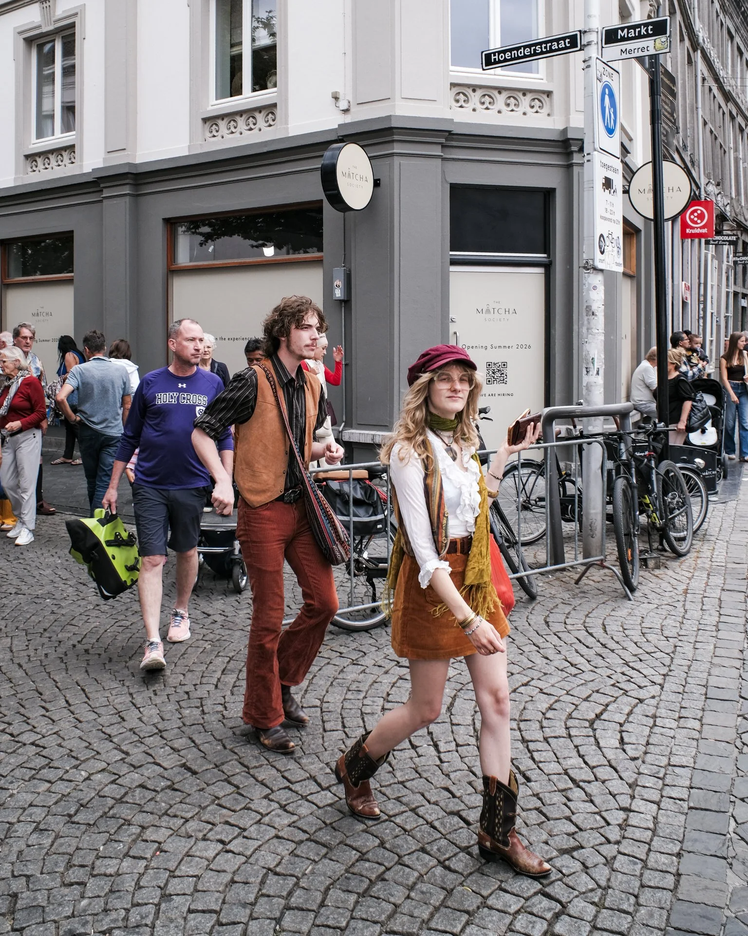 People crossing a cobblestone street at an intersection in an urban area, with storefronts and signage visible in the background.