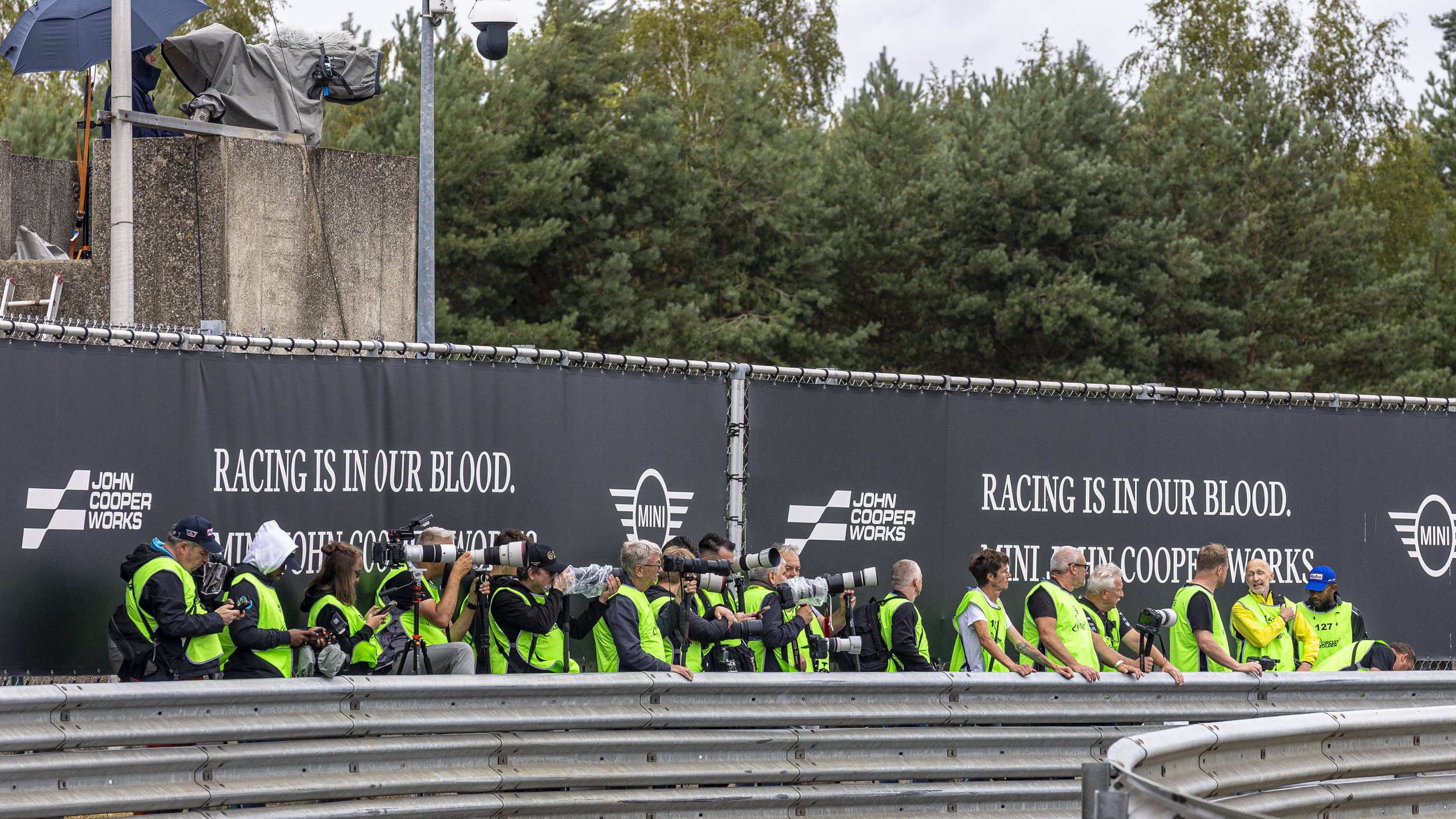 Group of people, some with cameras, wearing green safety vests at a racing event, with a black banner displaying the text 'RACING IS IN OUR BLOOD' and logos for John Cooper Works and MINI in the background, alongside trees.