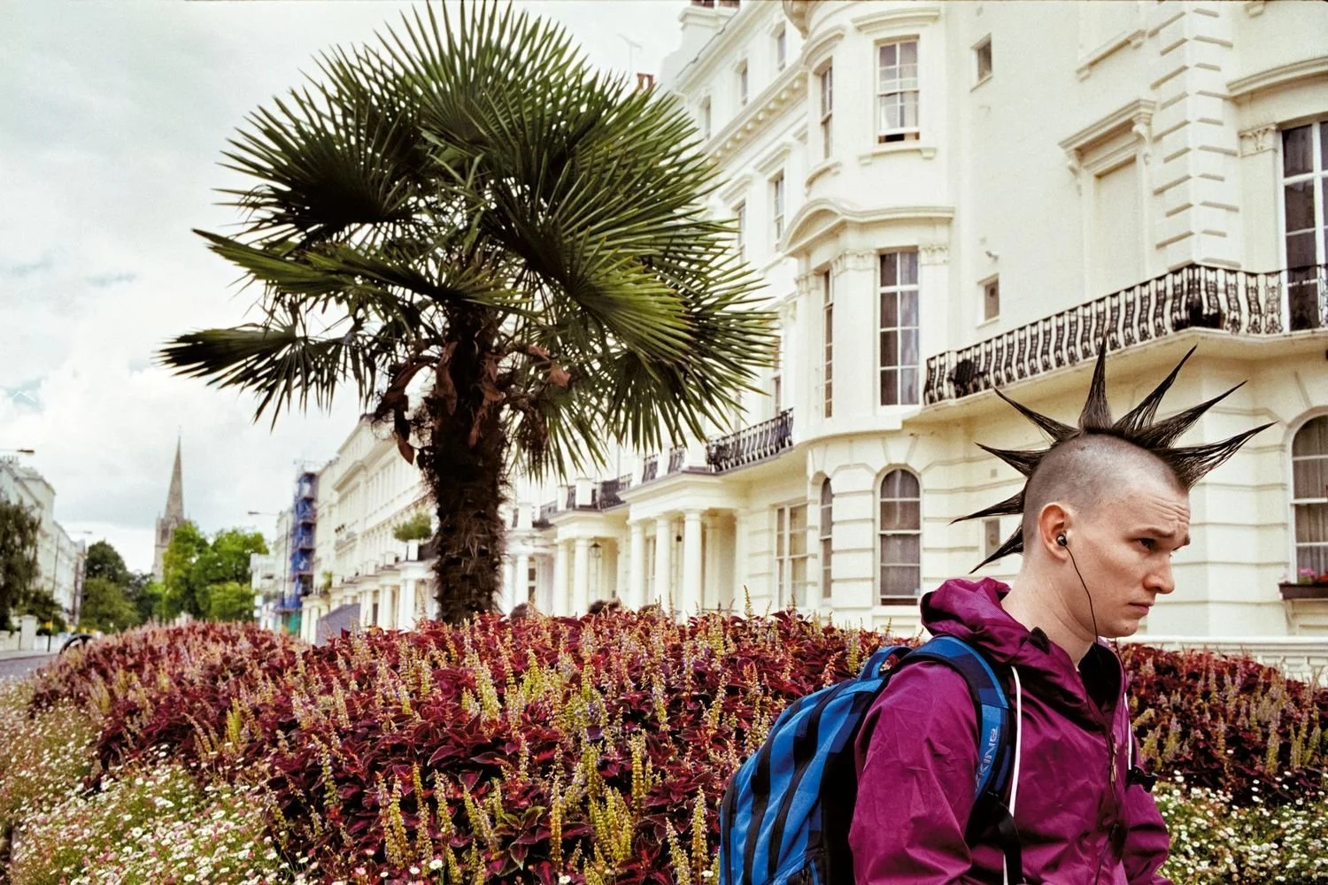 A person with a mohawk hairstyle made of spikes, wearing a purple jacket and a blue backpack, standing in front of a garden with a tall palm tree and white historic buildings with black wrought iron balconies.