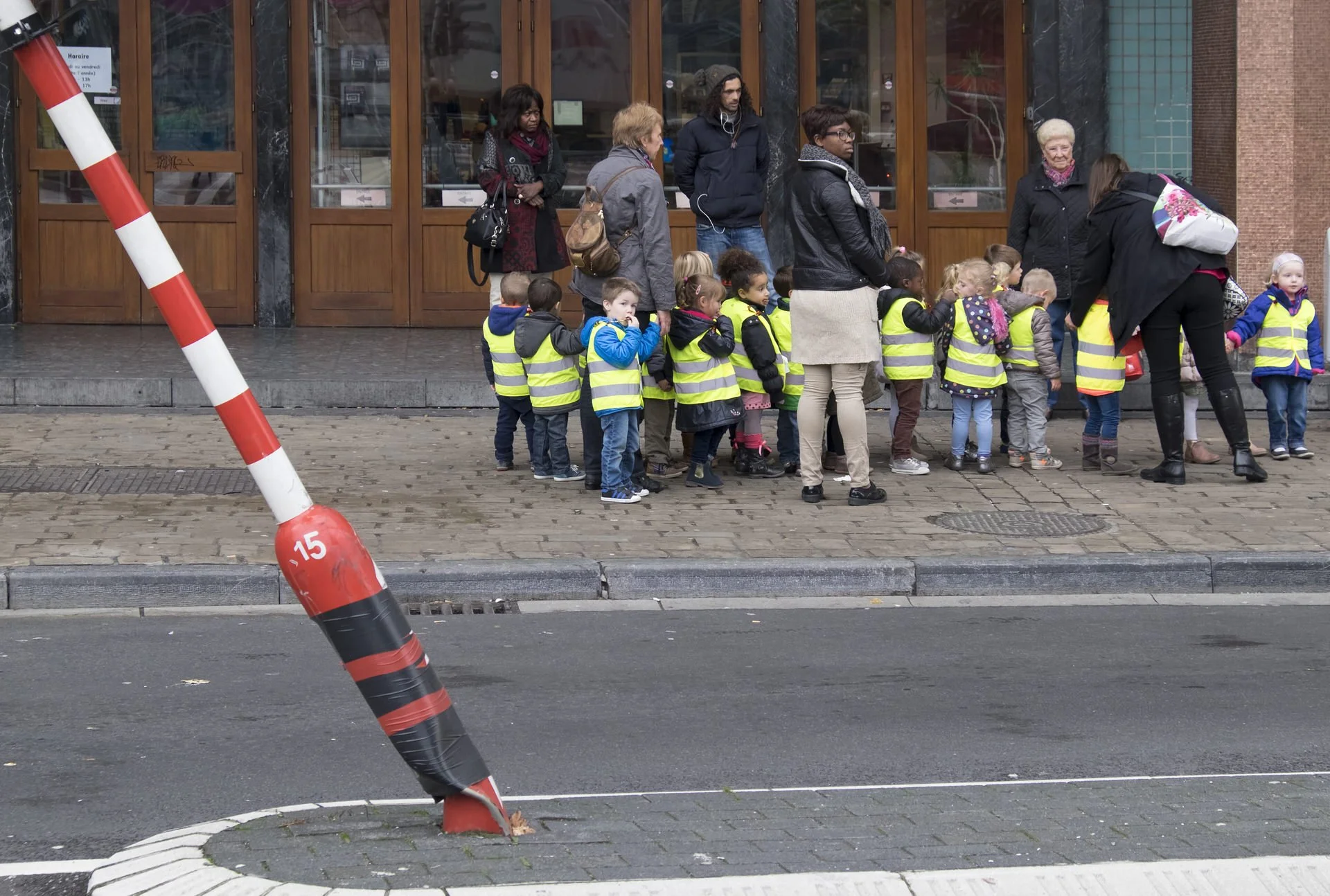 Line of young children wearing yellow reflective safety vests standing on the sidewalk in front of a building with glass and wooden doors, accompanied by adults also in reflective vests, some with backpacks, while a traffic bollard leans on the curb 