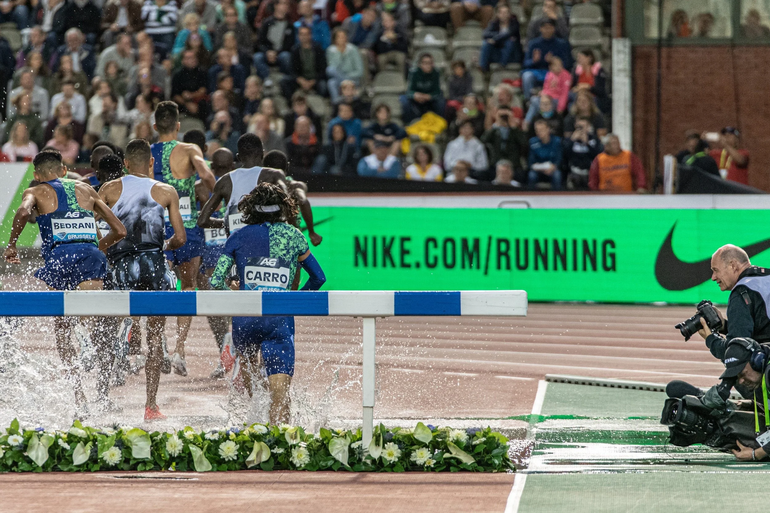 A group of athletes running on a track, crossing a water obstacle during a race, with a crowd of spectators watching in the stadium.