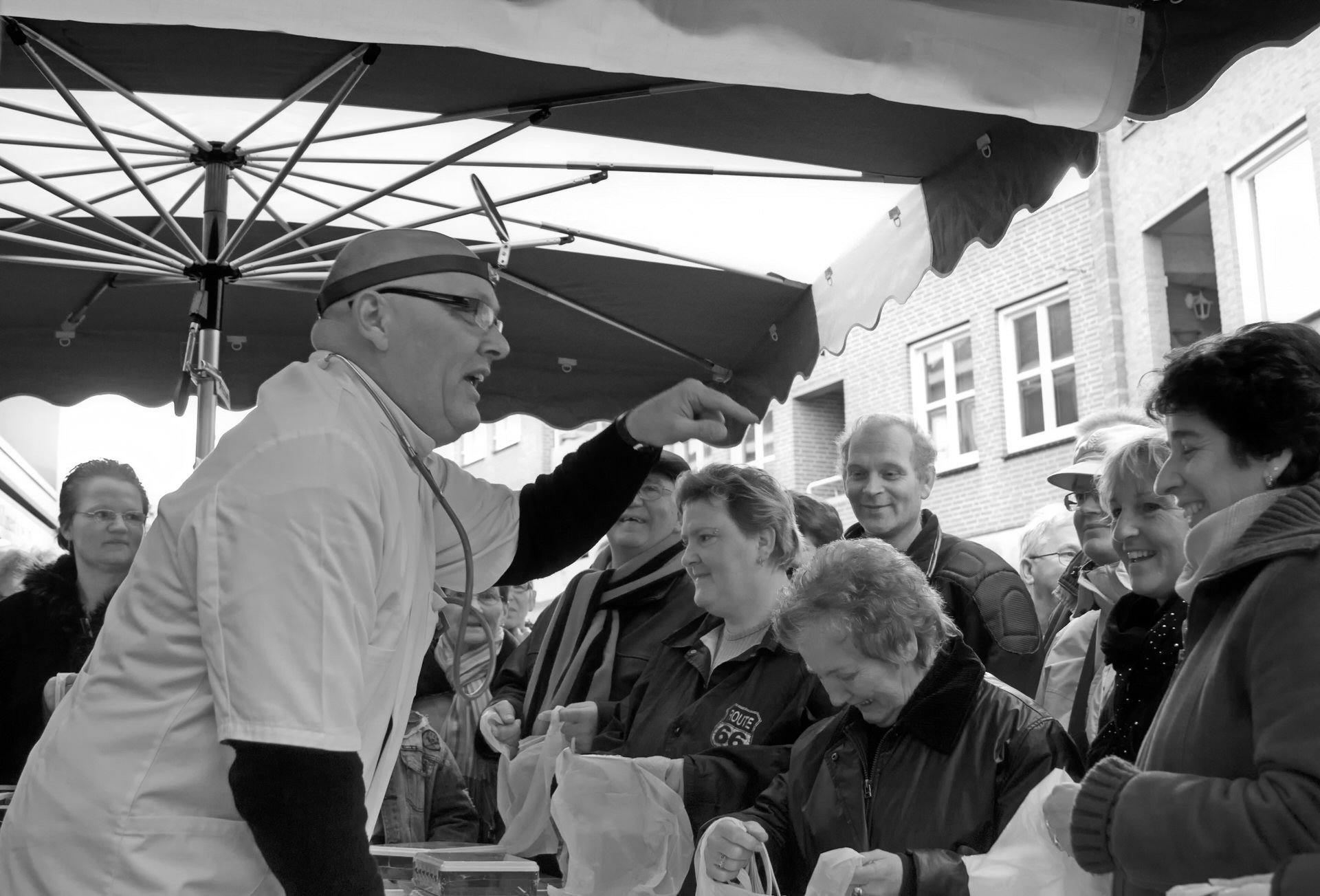 A black and white photo of a man in a white coat and glasses speaking to a crowd under a canopy. The crowd is gathered outdoors in front of a brick building, and some people are holding bags.