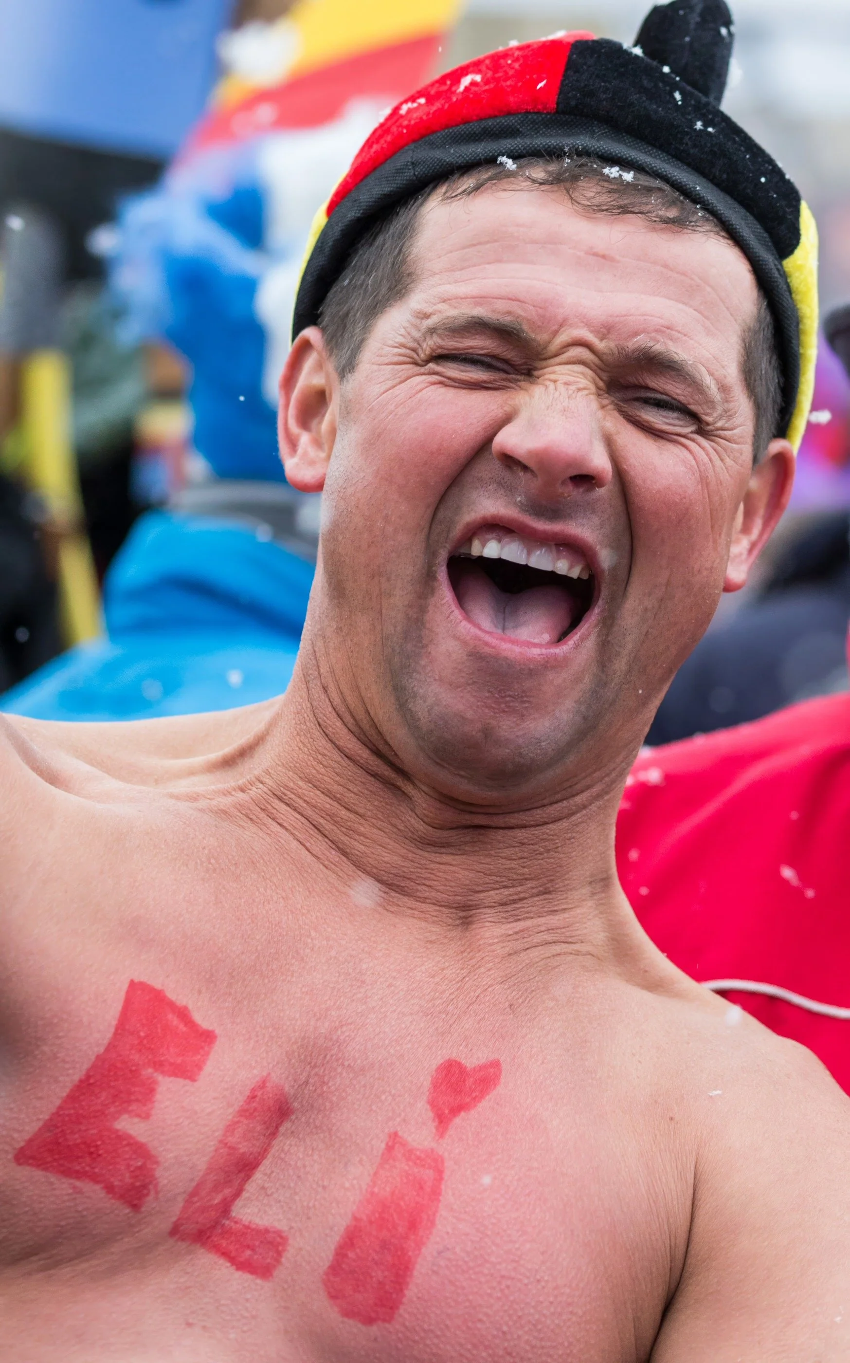 A man with short dark hair is laughing and shirtless, with red lipstick kisses on his chest and the word "LOVE" written in red on his skin.