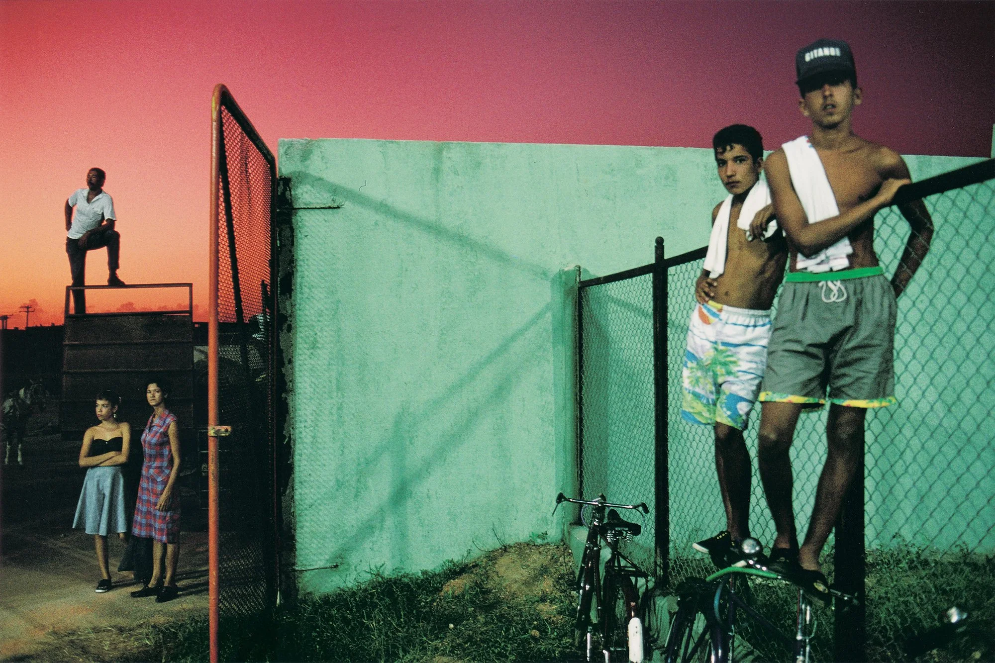 A group of young people, some shirtless and some in dresses, standing and leaning against a green wall and a chain-link fence. Two boys in the foreground are shirtless and wearing colorful swimwear, while three girls in the background are dressed in 