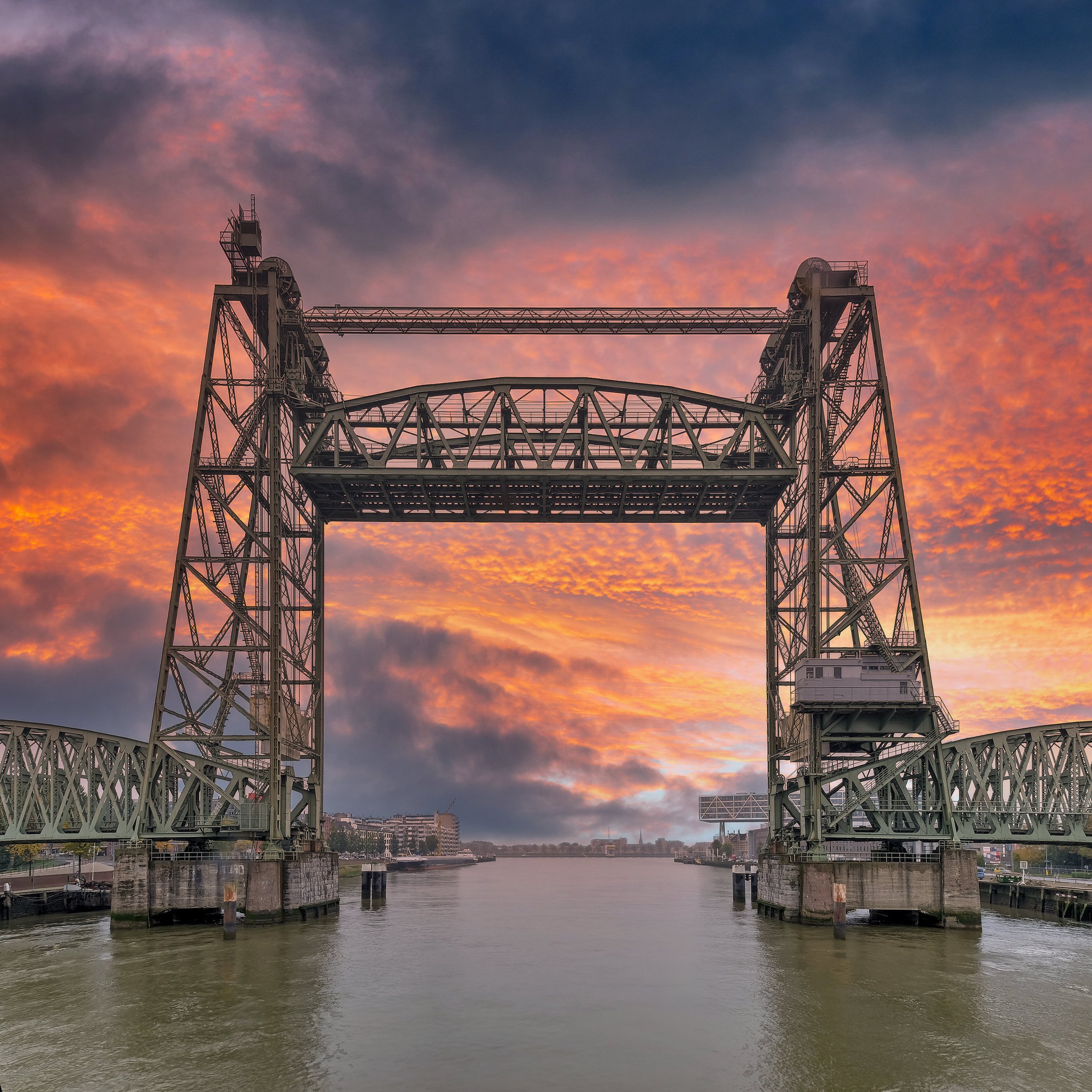 A steel bridge with towers and cables over a river at sunset, with a city skyline in the background.
