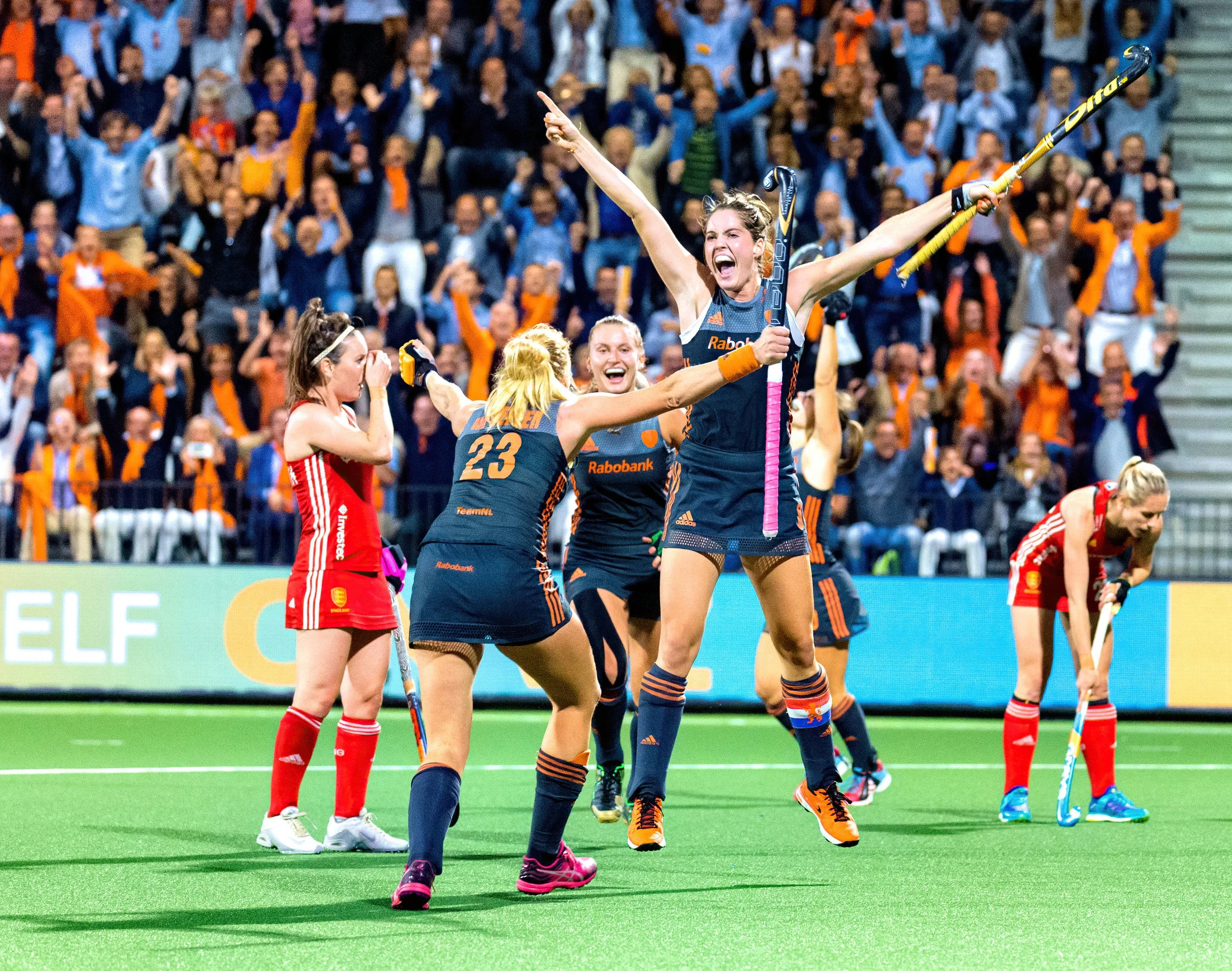 Women field hockey players celebrating on the field with a crowd in the background.