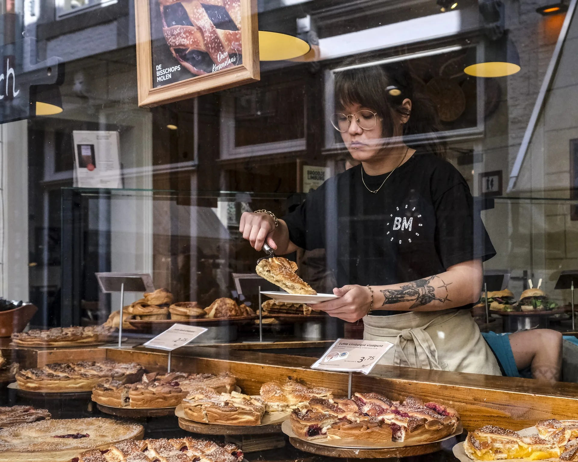 A woman with tattoos and glasses working behind a bakery display case filled with various pies and tarts, in a bakery storefront with signs in Dutch.