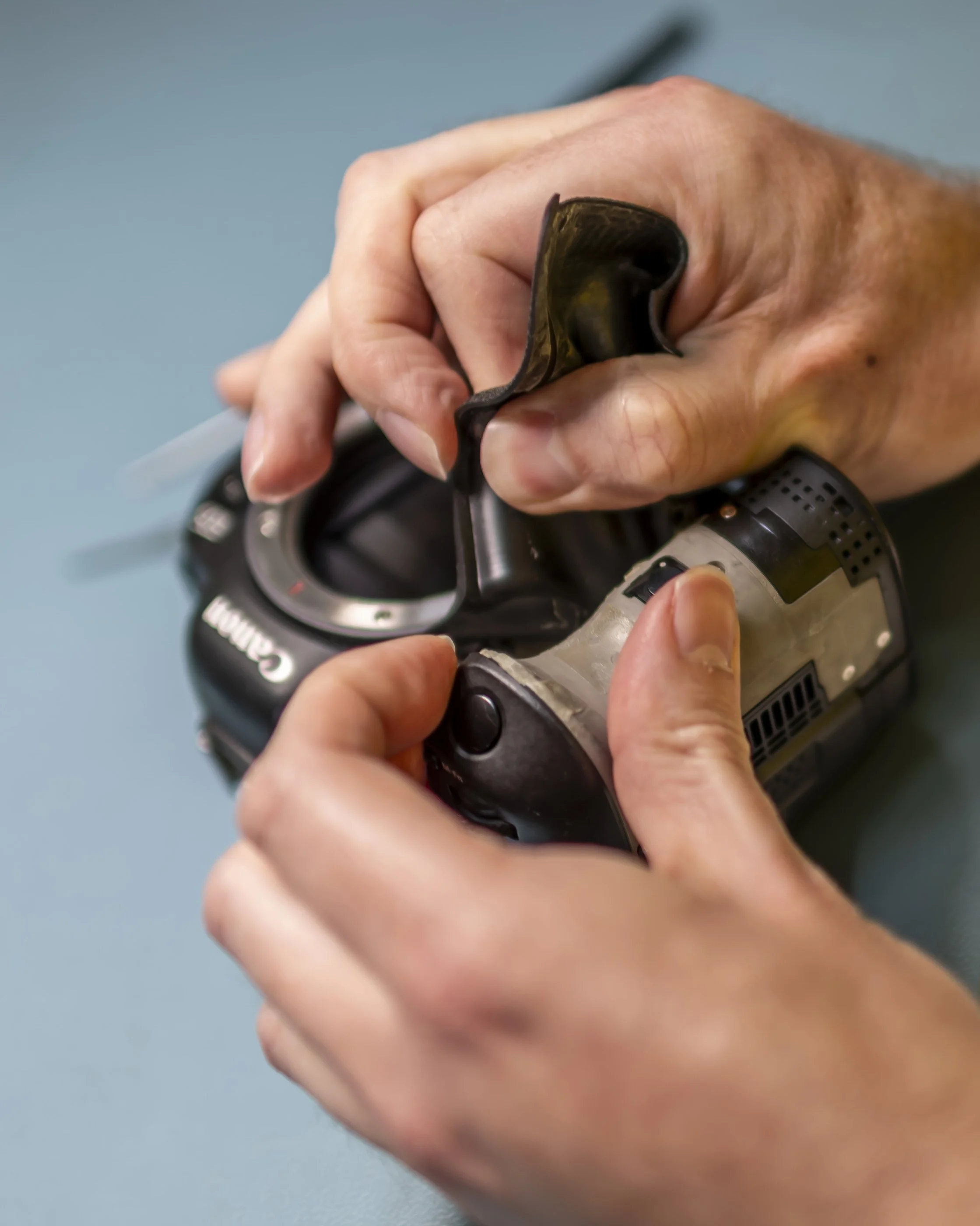 Close-up of a person's hand holding a drill, with a black rubber pad on their fingers, adjusting or repairing something. The background is a plain teal surface.