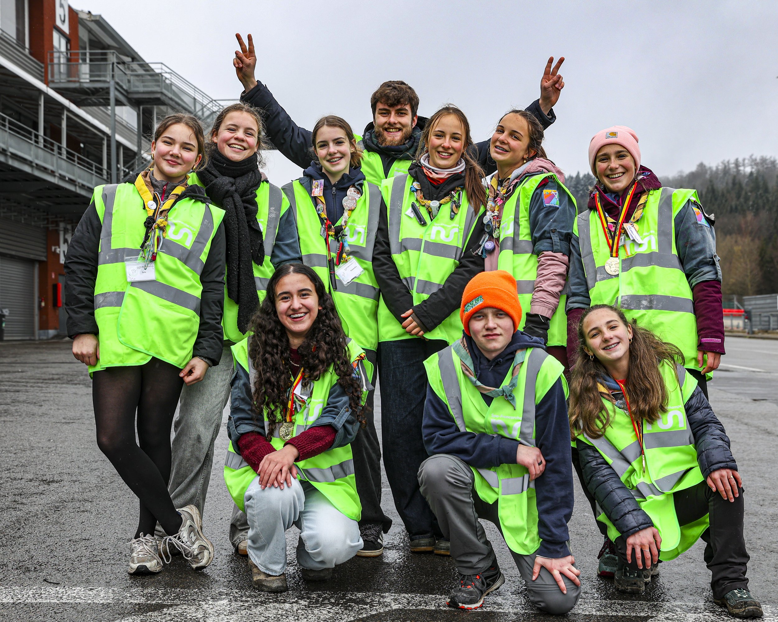 Group of children and a young man, all wearing bright green vests, posing outdoors on a cloudy day. Some children are smiling and kneeling, while the young man is standing with arms raised, making peace signs. They are at a location with metal struct