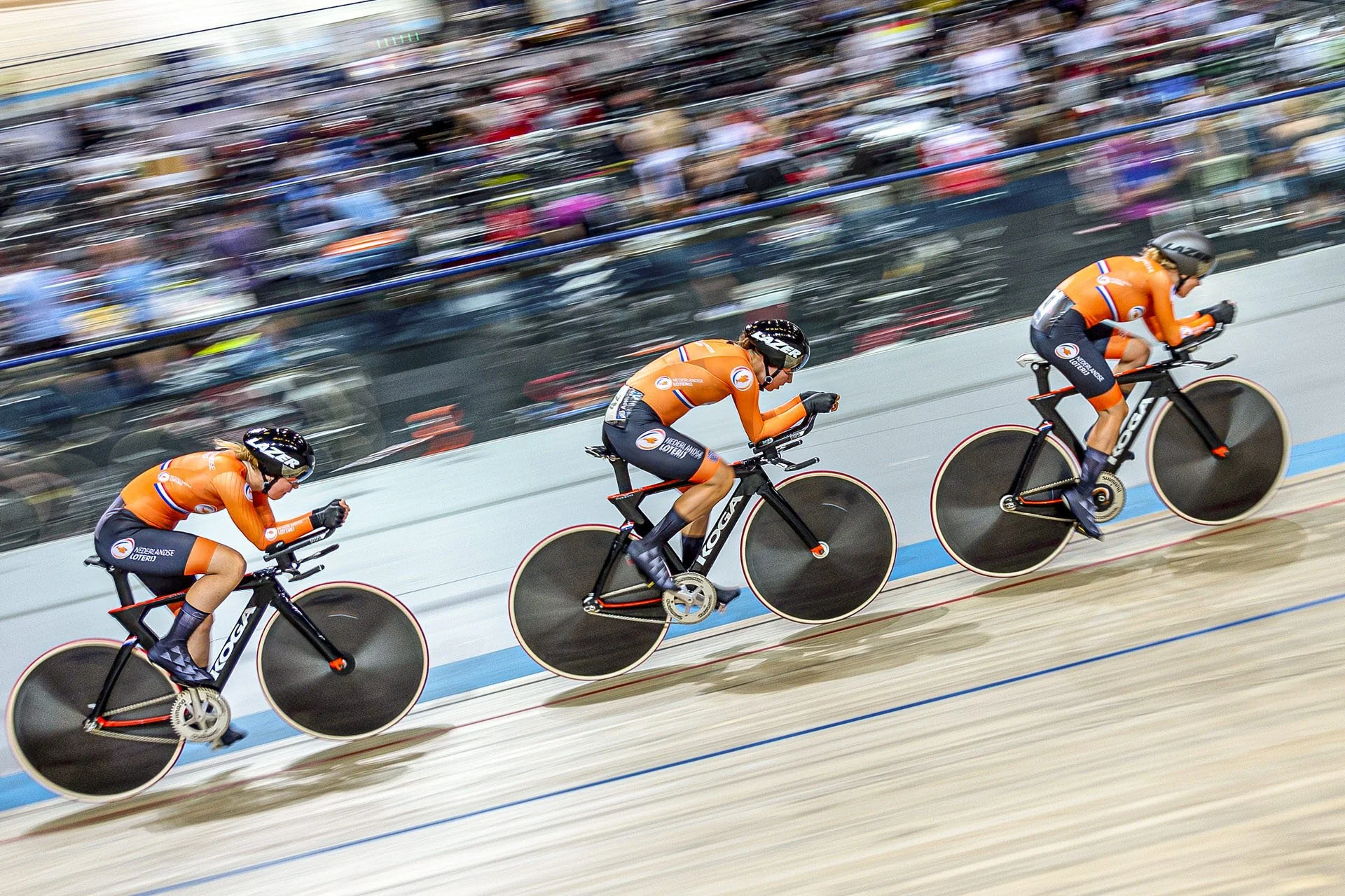Three cyclists in matching orange jerseys and black bikes racing on an indoor velodrome track with blurry audience in the background.