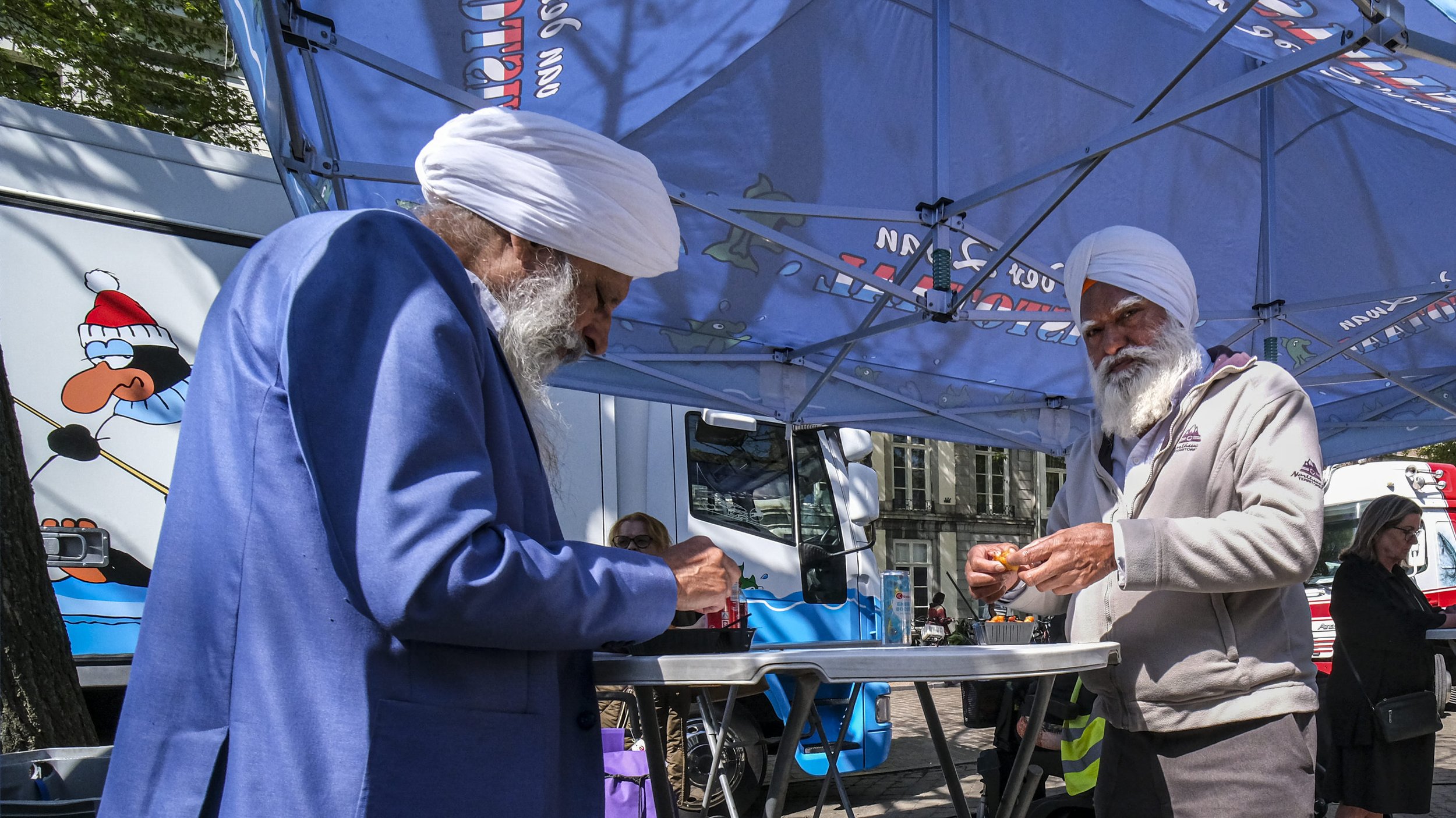 Two men with white turbans and beards are under a blue canopy at an outdoor event. They are focused on something in their hands, and there are other people and a truck in the background.