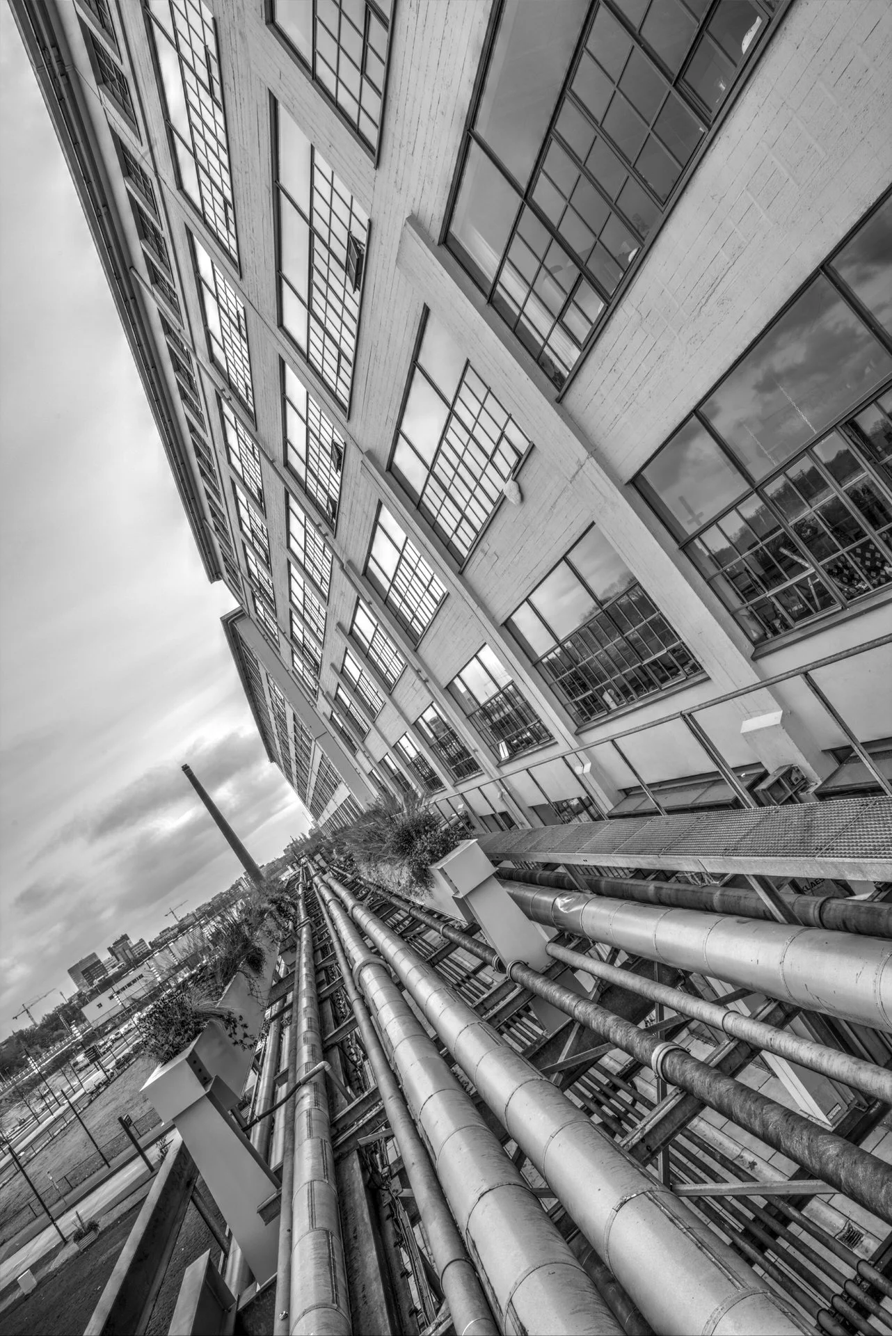 Black and white photo of a modern multi-story office or apartment building with numerous windows and reflective glass. Pipes run along the exterior lower part of the building, leading toward the horizon. The sky is cloudy, and city buildings are visi